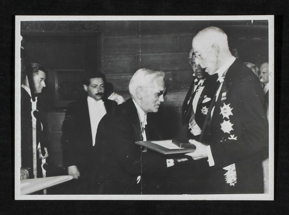 Black and white photo of formally dressed men, one handing a document or folder to another, in an ornate room with wood paneling.