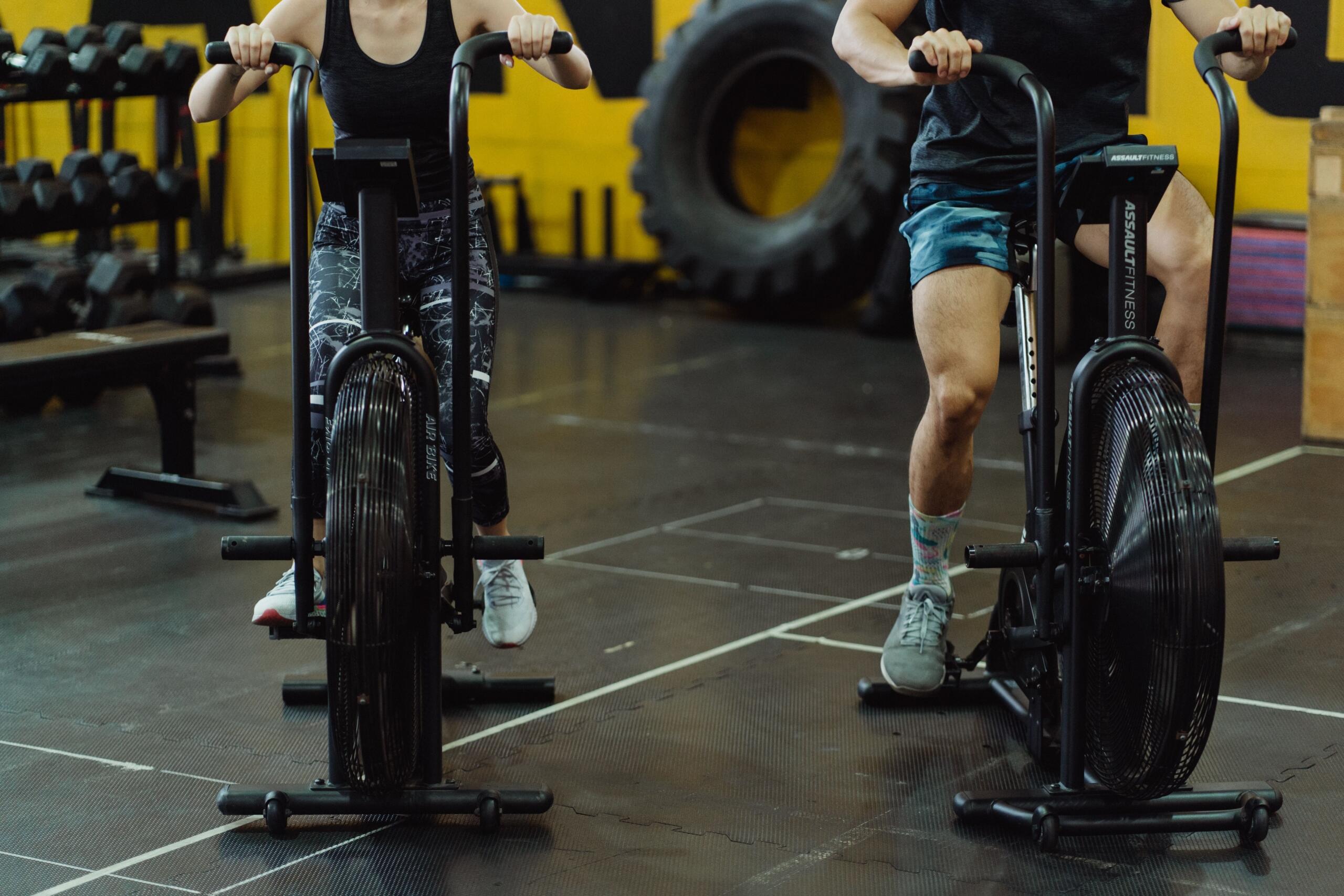 A group of people trains on air bikes in a gym, pedaling intensely during a high-energy cardio workout while surrounded by fitness equipment.