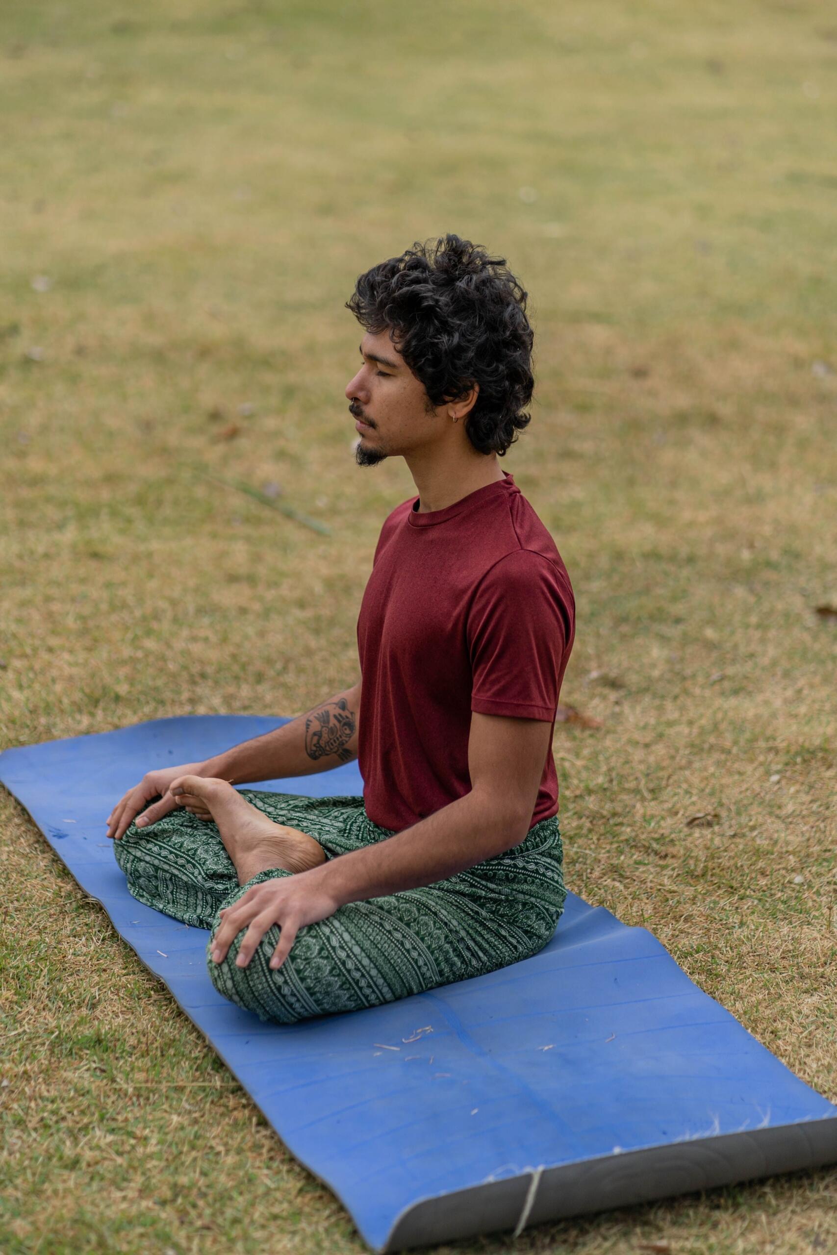 A man sits cross-legged on a yoga mat outdoors, meditating peacefully in a natural setting surrounded by grass and trees.