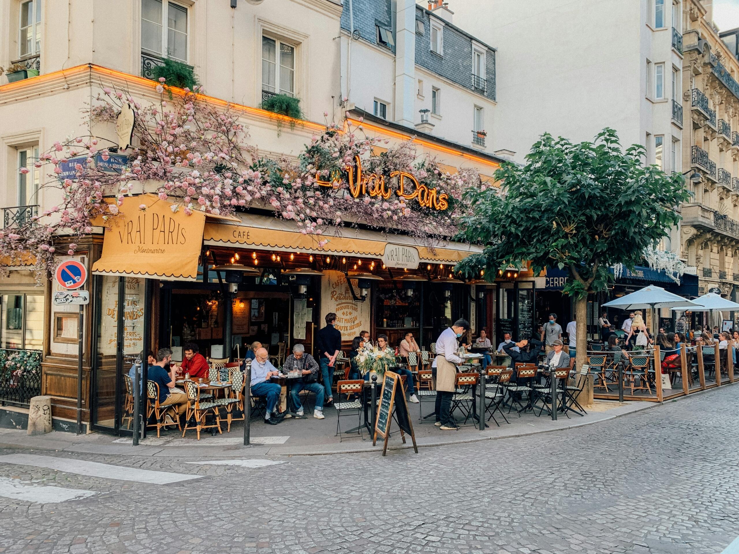 Bustling Parisian café named “Le Vrai Paris” with pink flowers decorating the awning, outdoor seating filled with people dining, and a waiter serving tables on a cobblestone street corner in Montmartre.