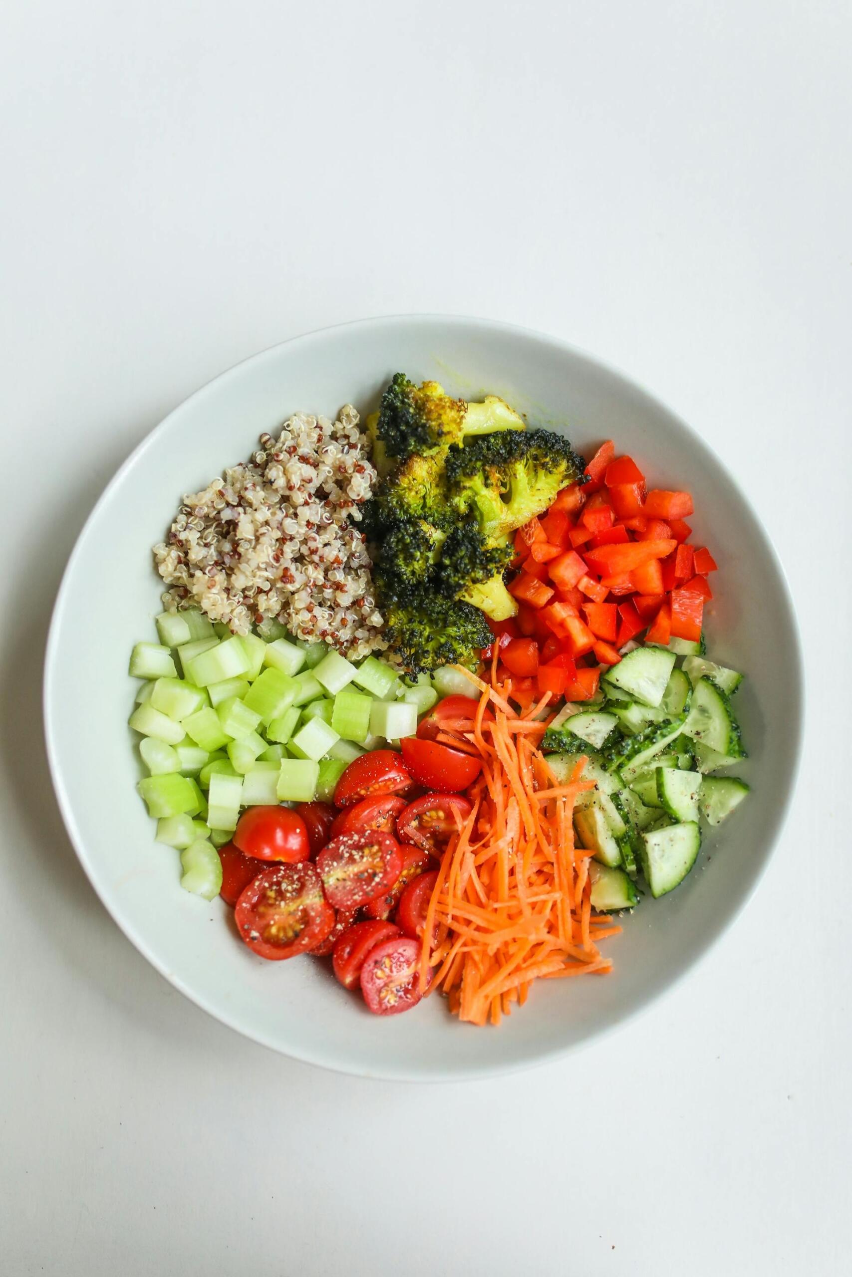 A vibrant bowl filled with quinoa, broccoli, diced red peppers, cucumber, celery, cherry tomatoes, and shredded carrots.