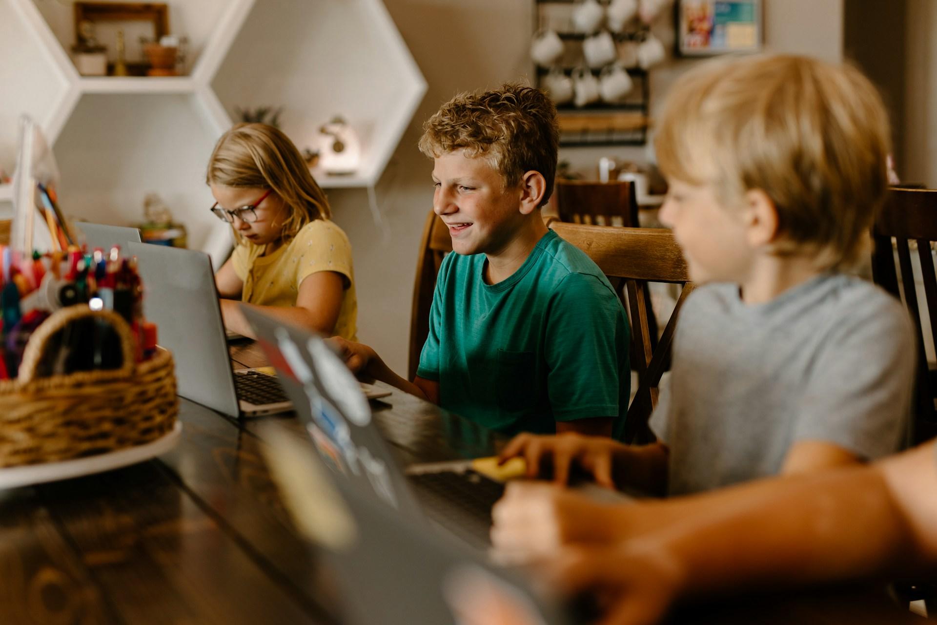 Three children engaged in activities at a table with laptops and stationery.