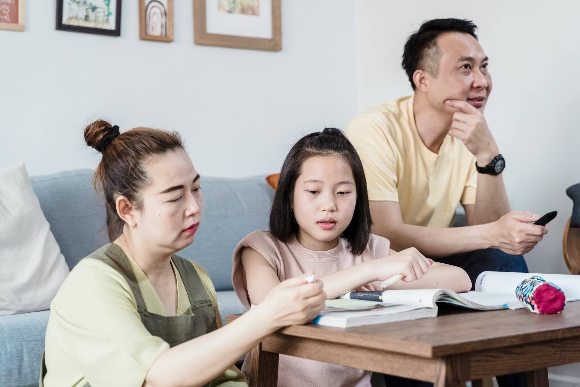 Parents sitting with their daughter at a table, helping her with homework in a cozy living room, illustrating parental support in a student’s learning process.