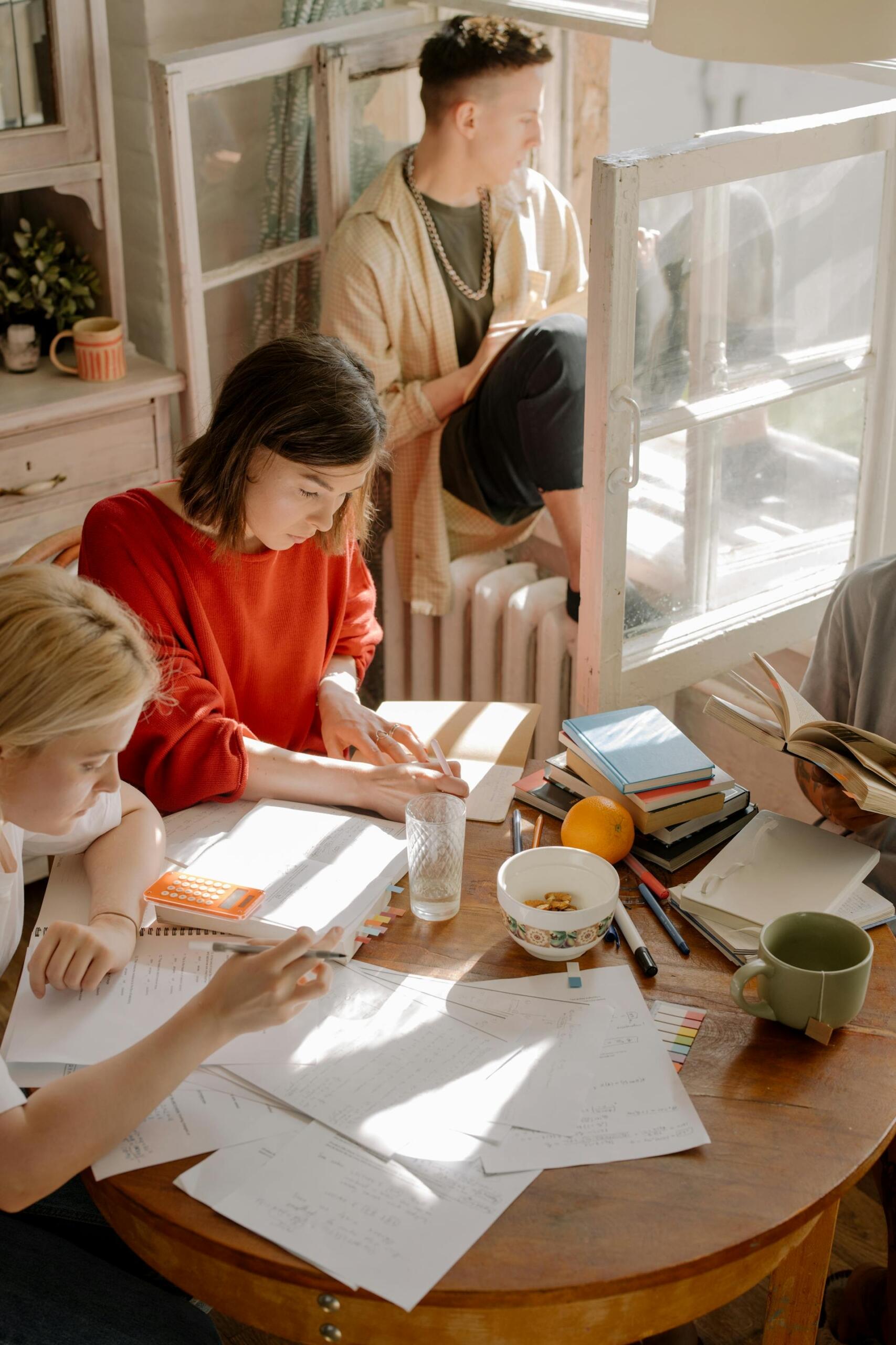 Group of students studying together at a wooden table filled with notebooks, papers, and books in a bright, cozy room with natural sunlight coming through the window.