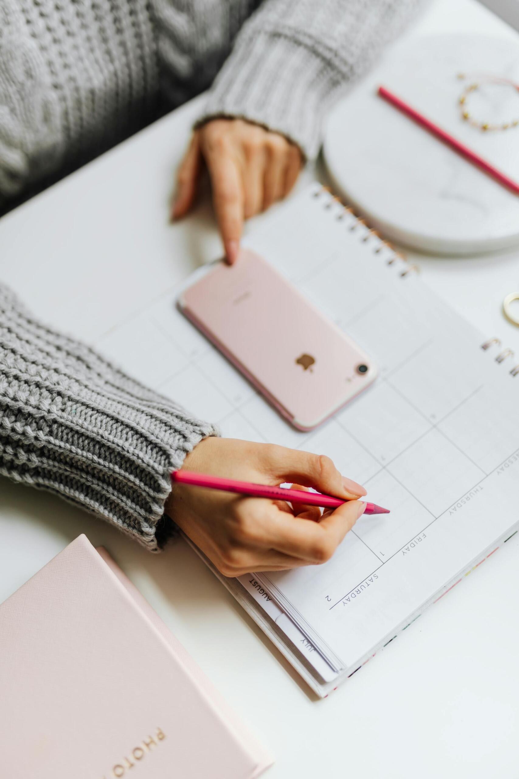 Close-up of a person planning their schedule with a pink pencil on a calendar, next to a smartphone and notebook on a white desk, representing time management and study organization.
