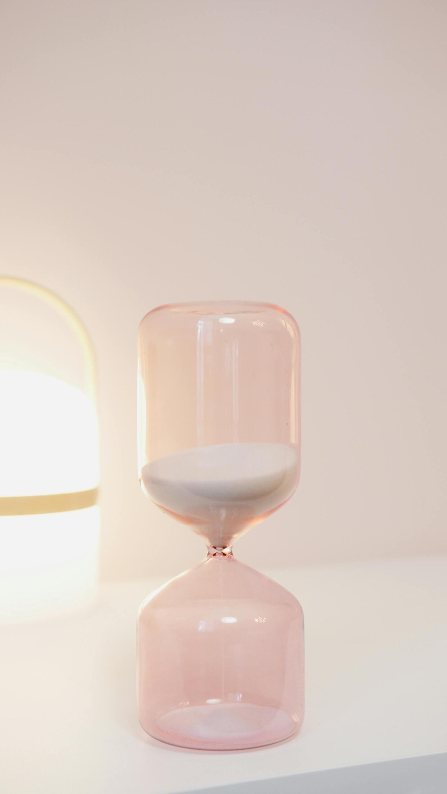 A modern pink glass hourglass with white sand placed on a white surface, symbolizing time management and focus in a calm, minimalist setting.