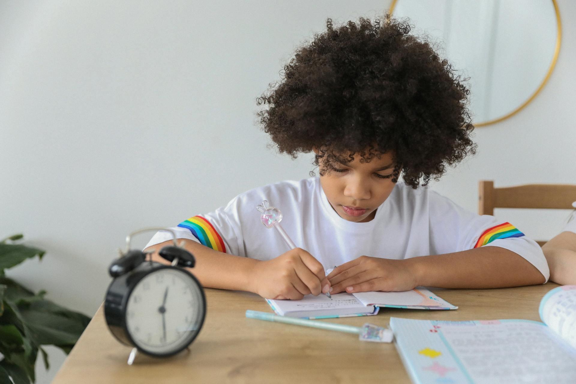A child sitting at a wooden desk writing in a notebook, with a black alarm clock and school supplies nearby, focusing on homework in a bright, tidy study space.