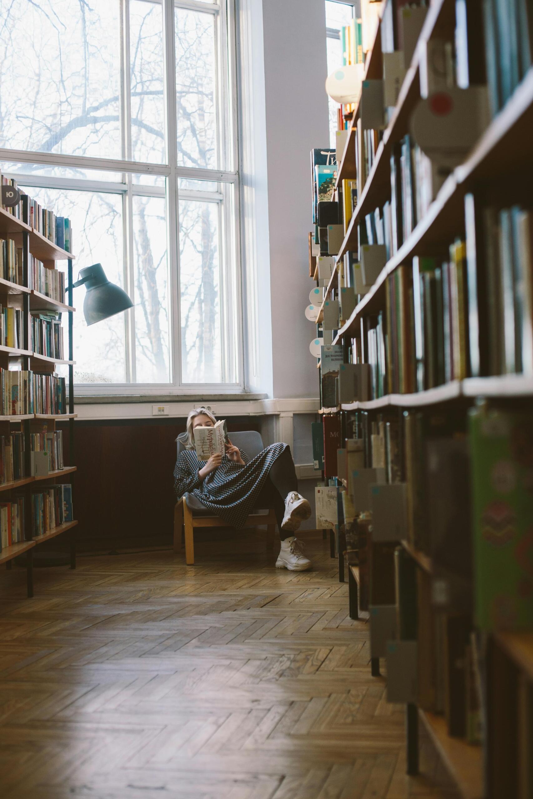 A woman sits in a library reading a book under a warm desk lamp, with rows of bookshelves stretching out in front of her.