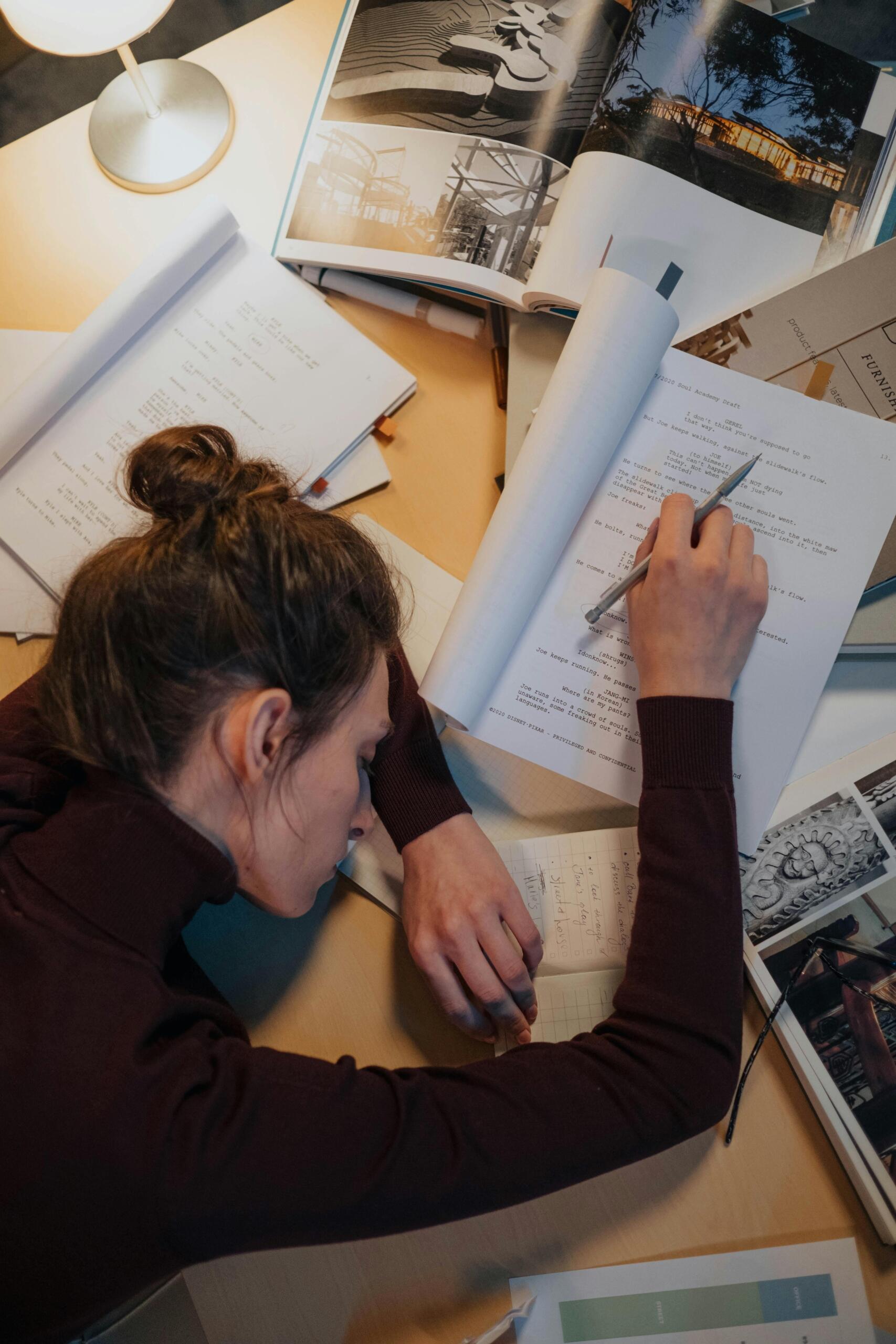 A student rests her head on her desk, having fallen asleep over her schoolwork, with papers and a notebook spread out in front of her.