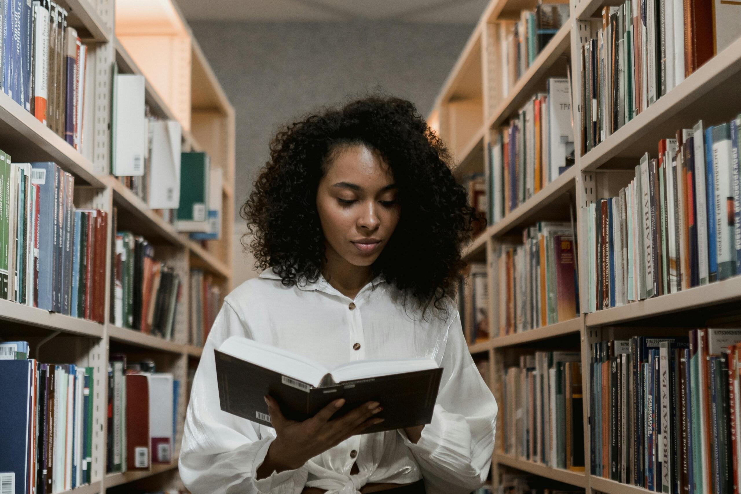 A woman stands in a quiet library aisle, reading a book while surrounded by tall shelves filled with neatly arranged books.