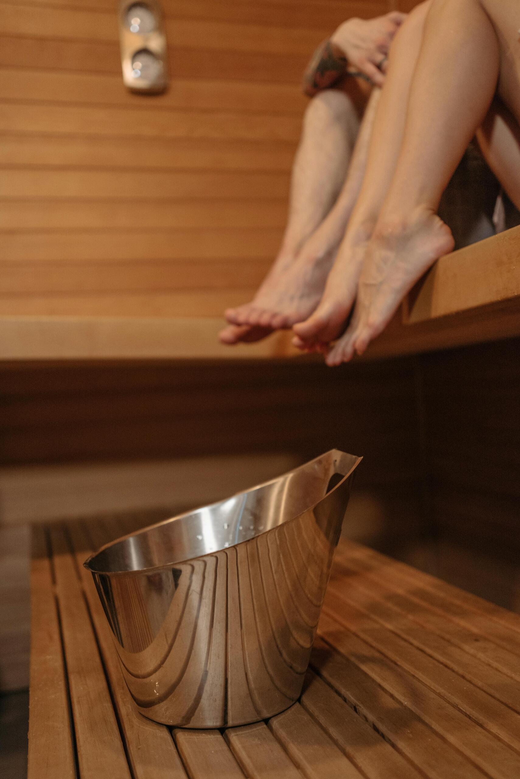 A close-up of bare feet resting on a wooden bench inside a sauna, surrounded by warm wooden panels and gentle ambient lighting.