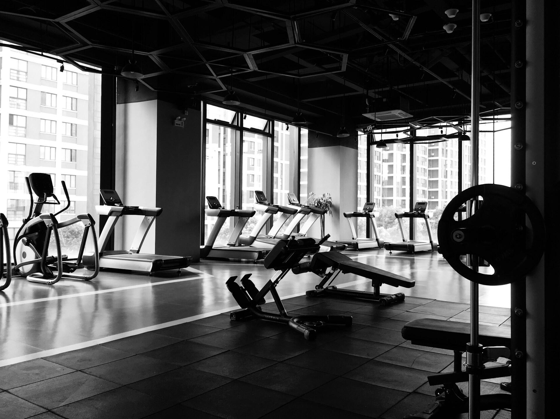 Black and white photo of a modern gym interior with treadmills, exercise bikes, weight bench, and large windows showing buildings outside.