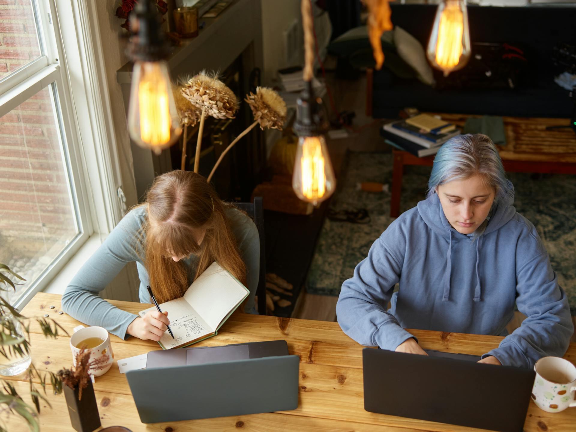Two individuals work at a wooden table, one writing in a notebook and the other using a laptop, with warm lighting and cozy decor.