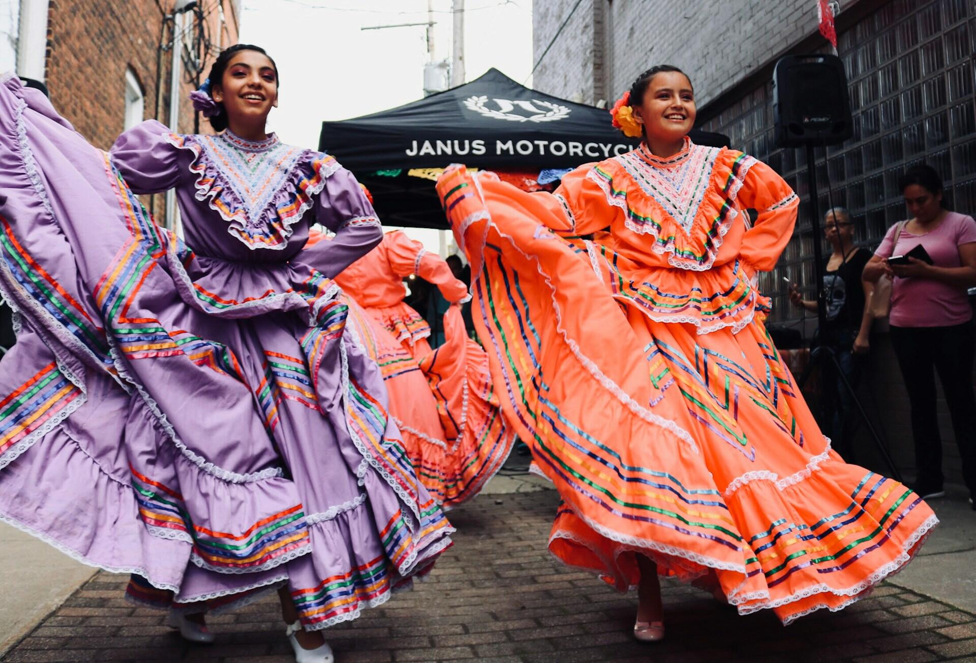 Two dancers in vibrant traditional dresses, one purple and one orange, perform gracefully in a lively street setting.