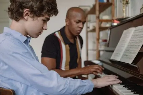 A young boy learning to play the piano with his tutor by his side.