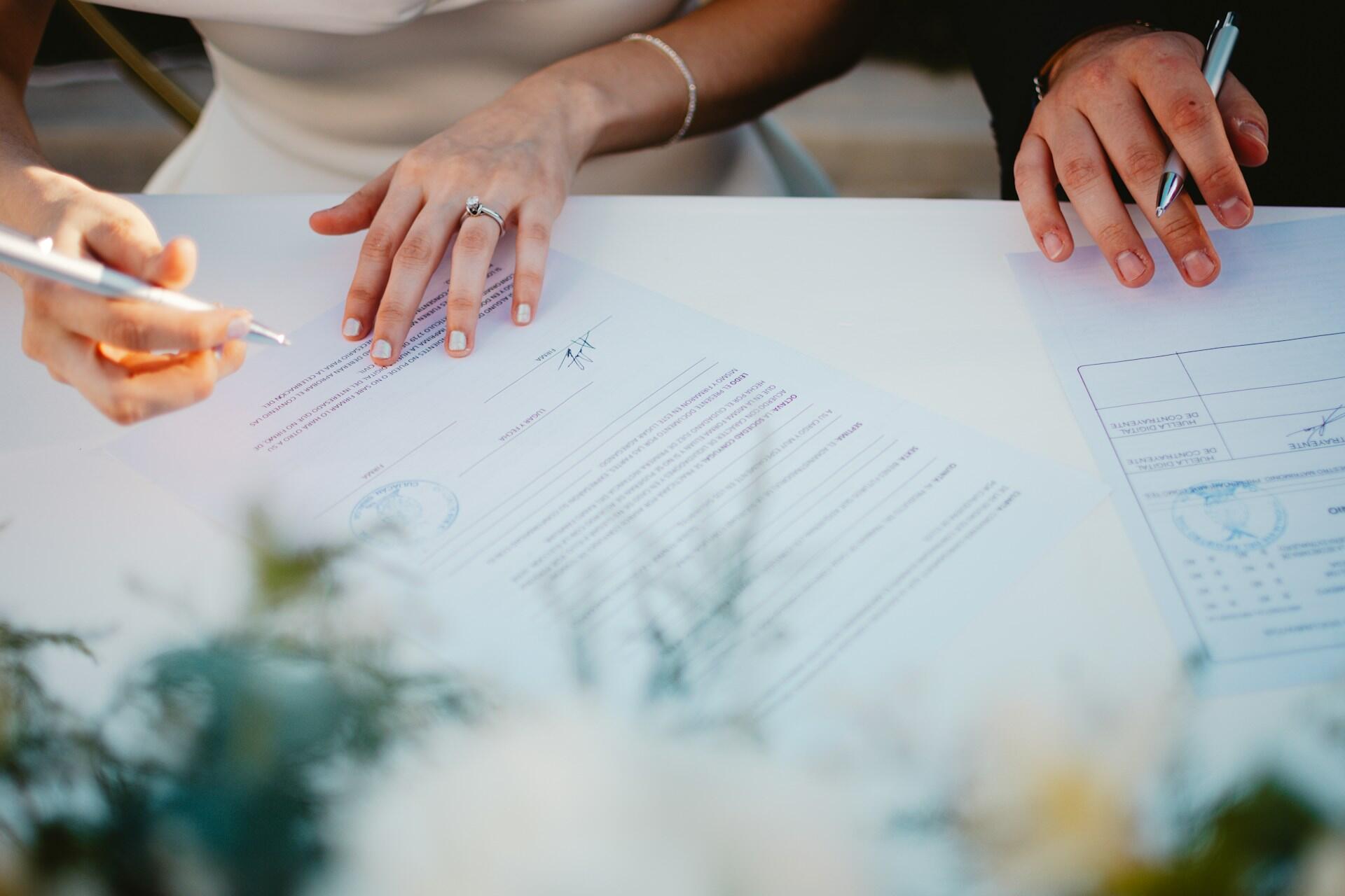 Close-up of a couple signing marriage documents at a table, with a bouquet blurred in the foreground.