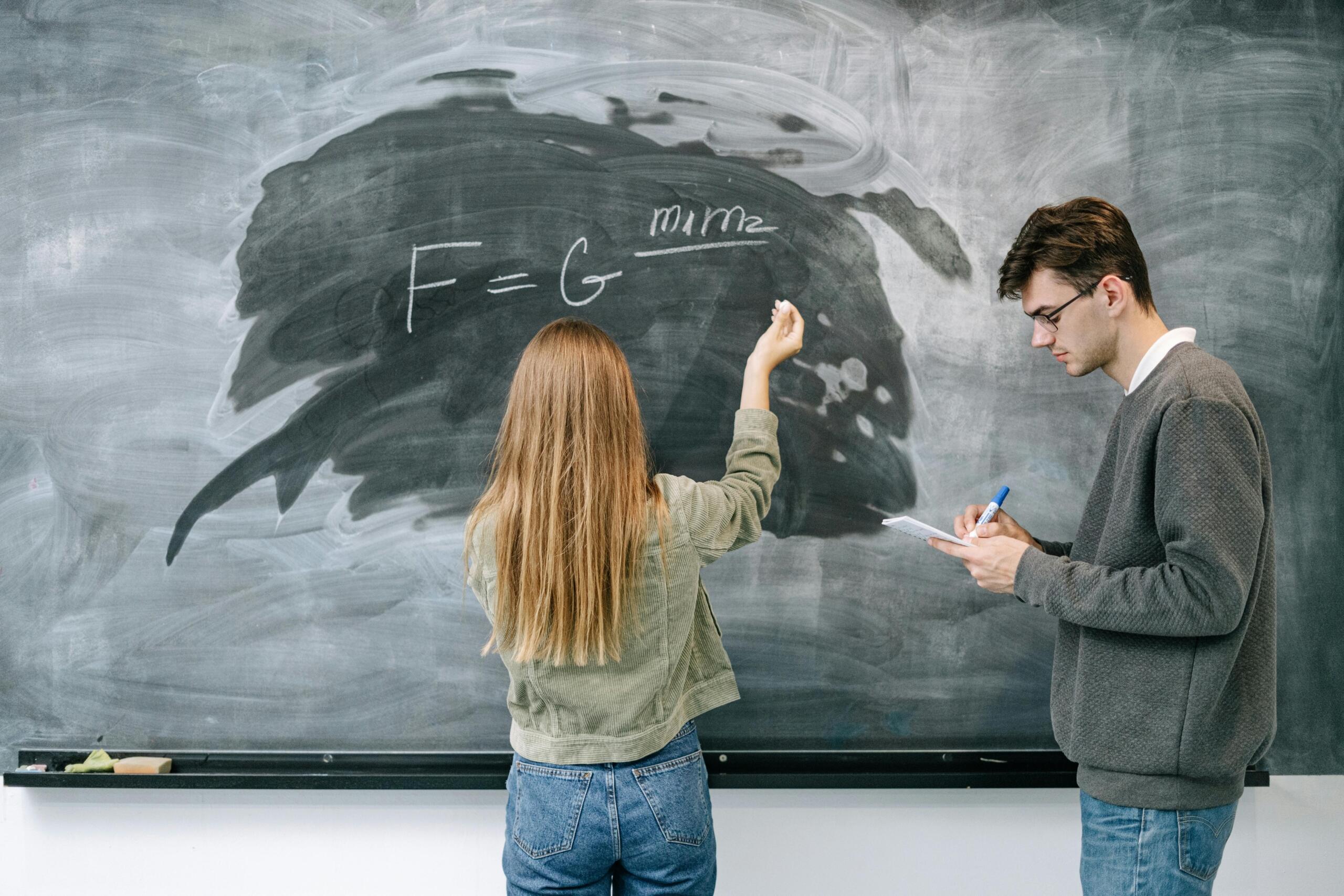 A teacher stands beside a blackboard covered with mathematical equations while helping a student solve a problem. The student looks attentively at the board as the teacher points to part of the equation during the explanation.