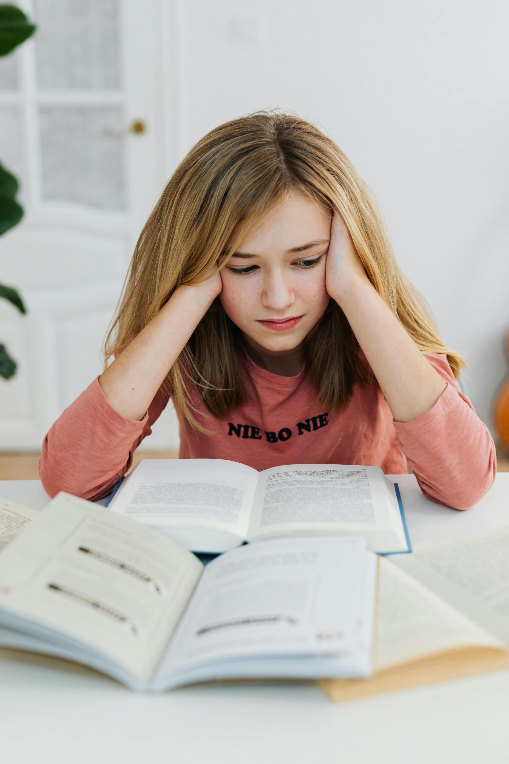 A teenage girl sits at a desk surrounded by textbooks, notes, and a laptop while studying. She rests her head in one hand and looks worried and overwhelmed as she reviews her work, suggesting stress or frustration with a challenging assignment or upcoming exam. Source: Karol G.