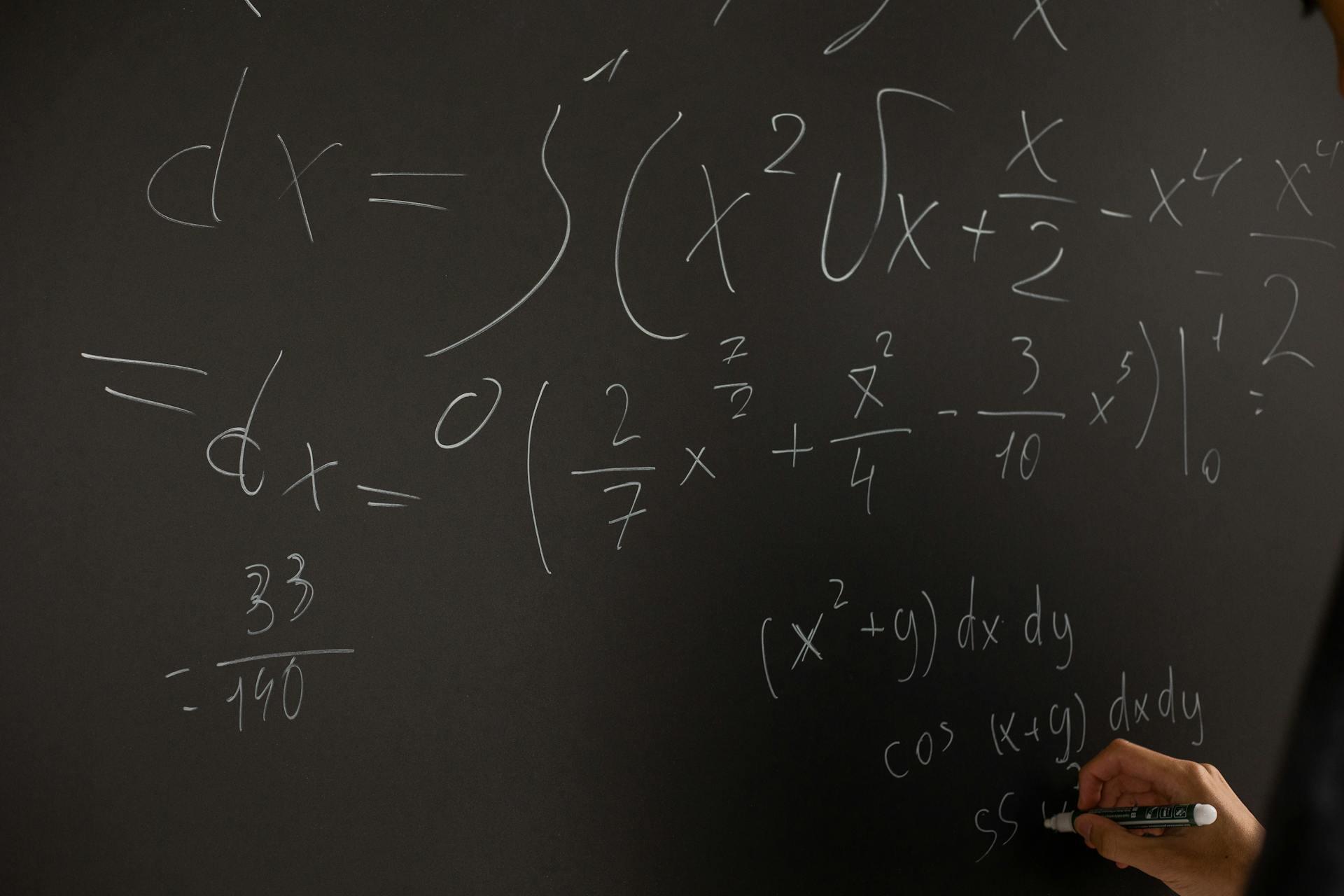 A person writes complex math equations in white chalk on a dark blackboard, including integrals and derivatives.