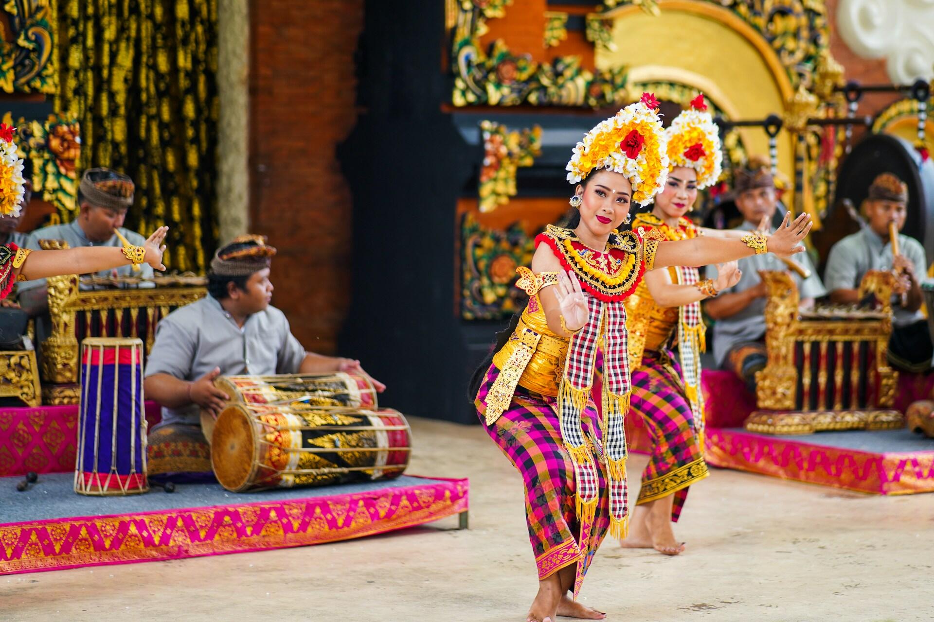 Two dancers in vibrant traditional attire perform on stage, accompanied by musicians playing intricate instruments in the background.