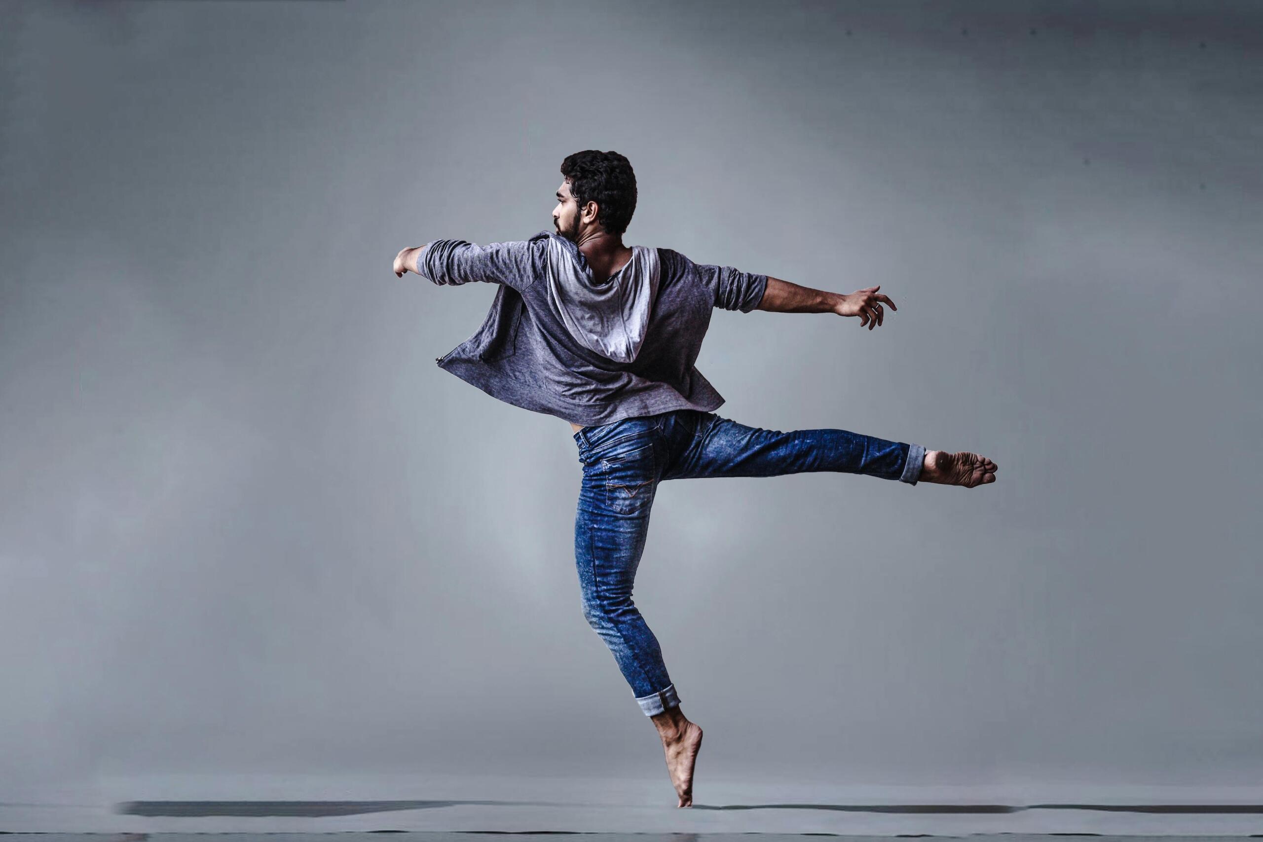 A man wearing denim and a hooded sweater dances in front of a gray background.