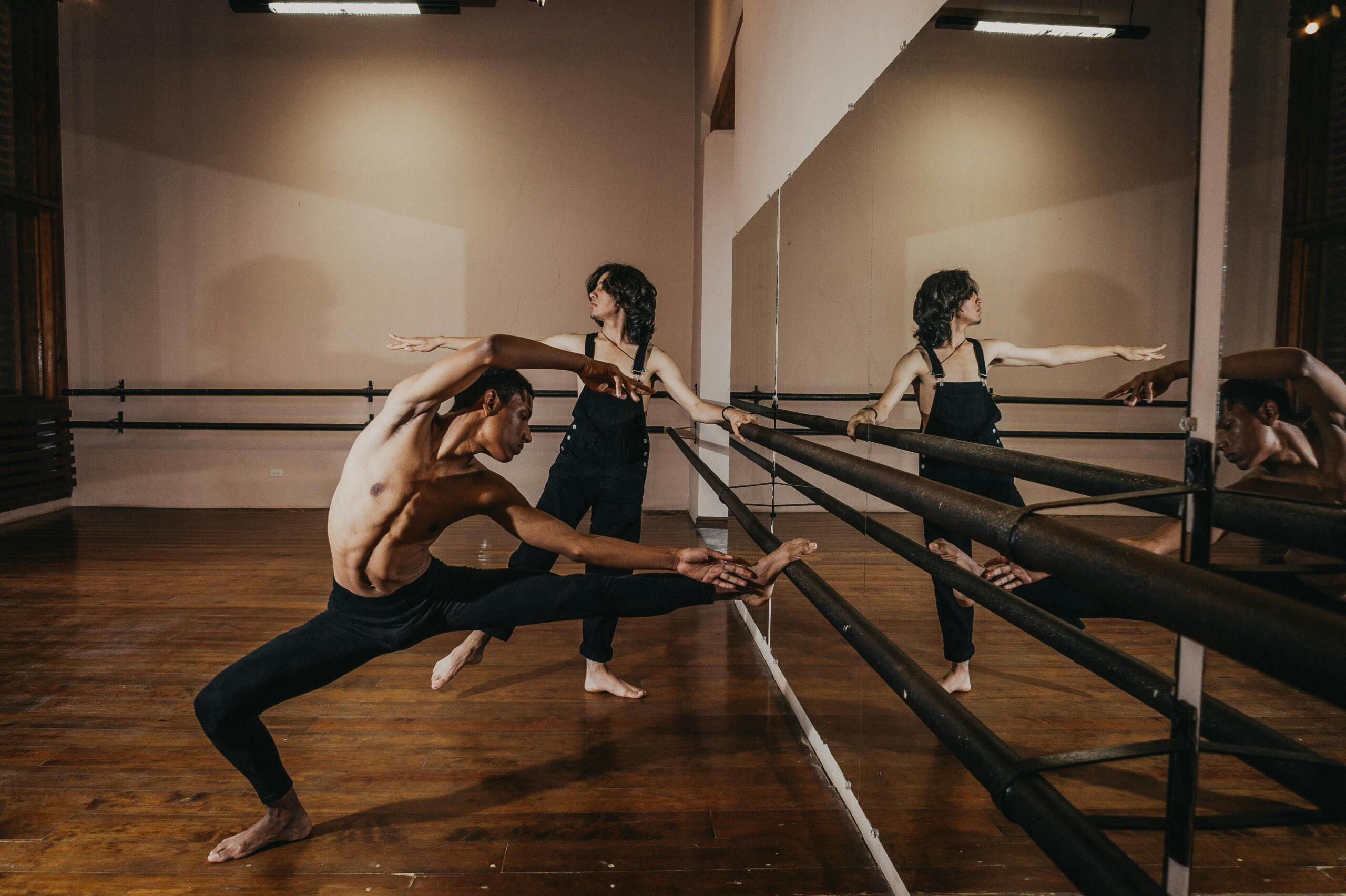 A man stretches at a barre in a dance studio. Another dancer in overalls dances behind him.