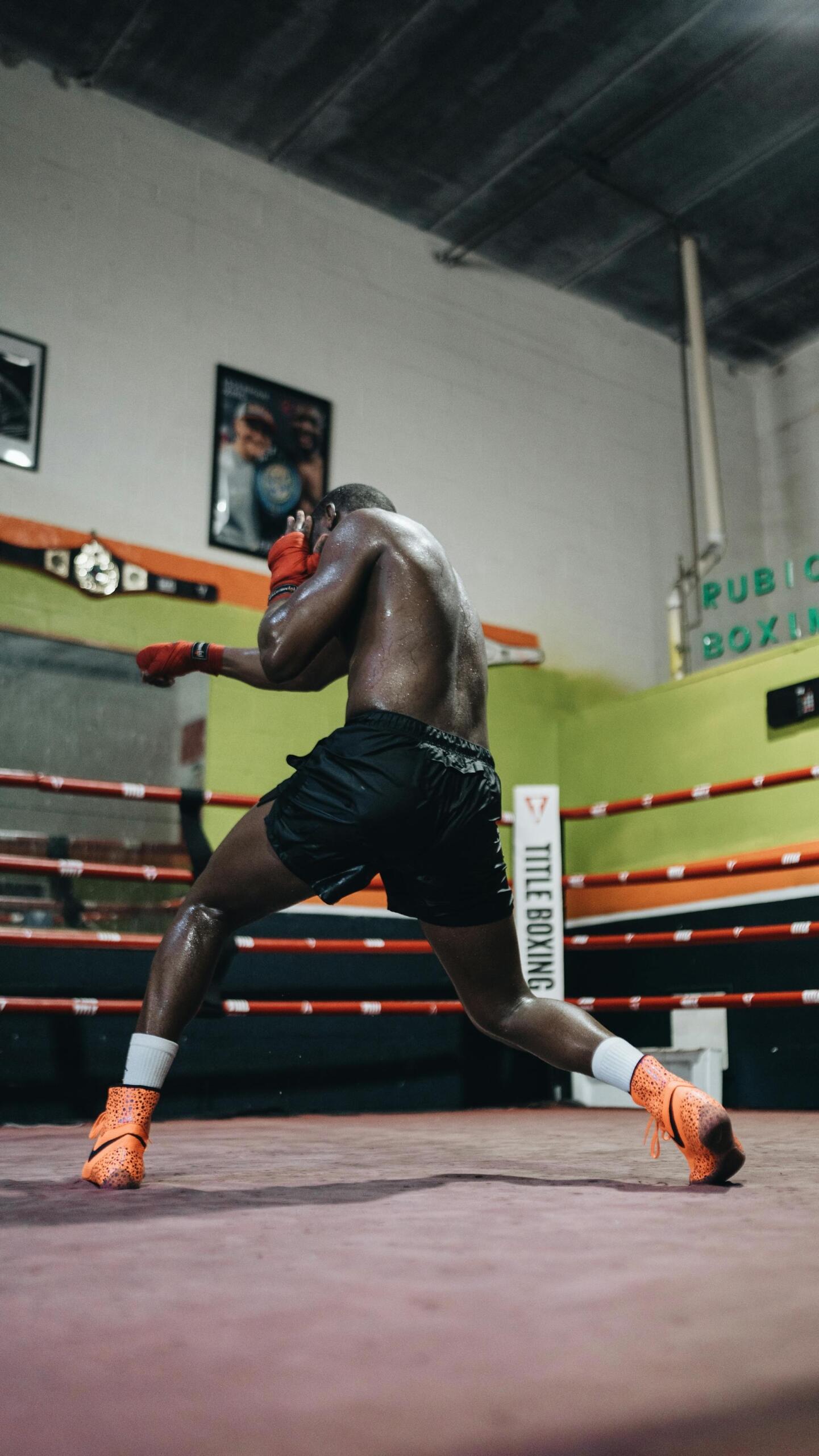 A shirtless boxer with red gloves trains in a ring, focusing on footwork. Vibrant orange shoes contrast against the gym's muted tones.