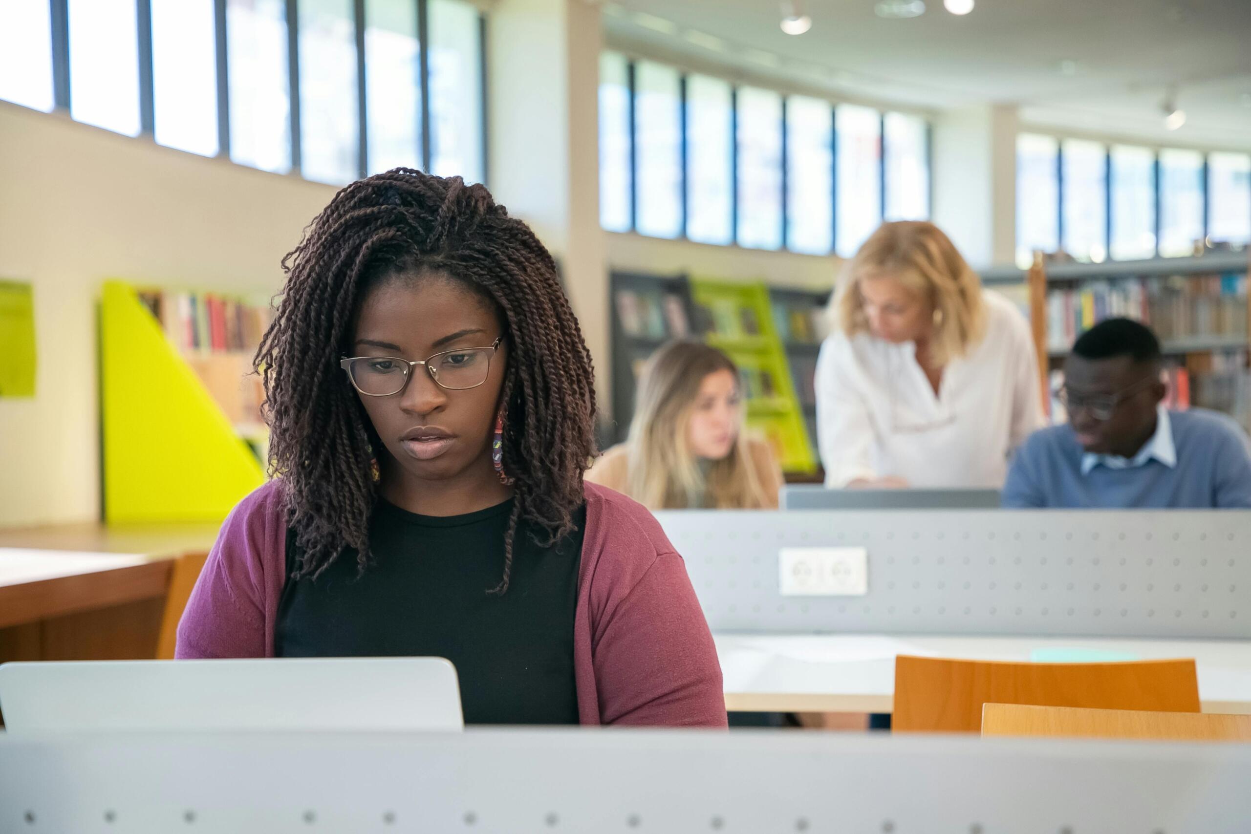 a woman looks at a laptop in a library. Two other students can be seen behind her.