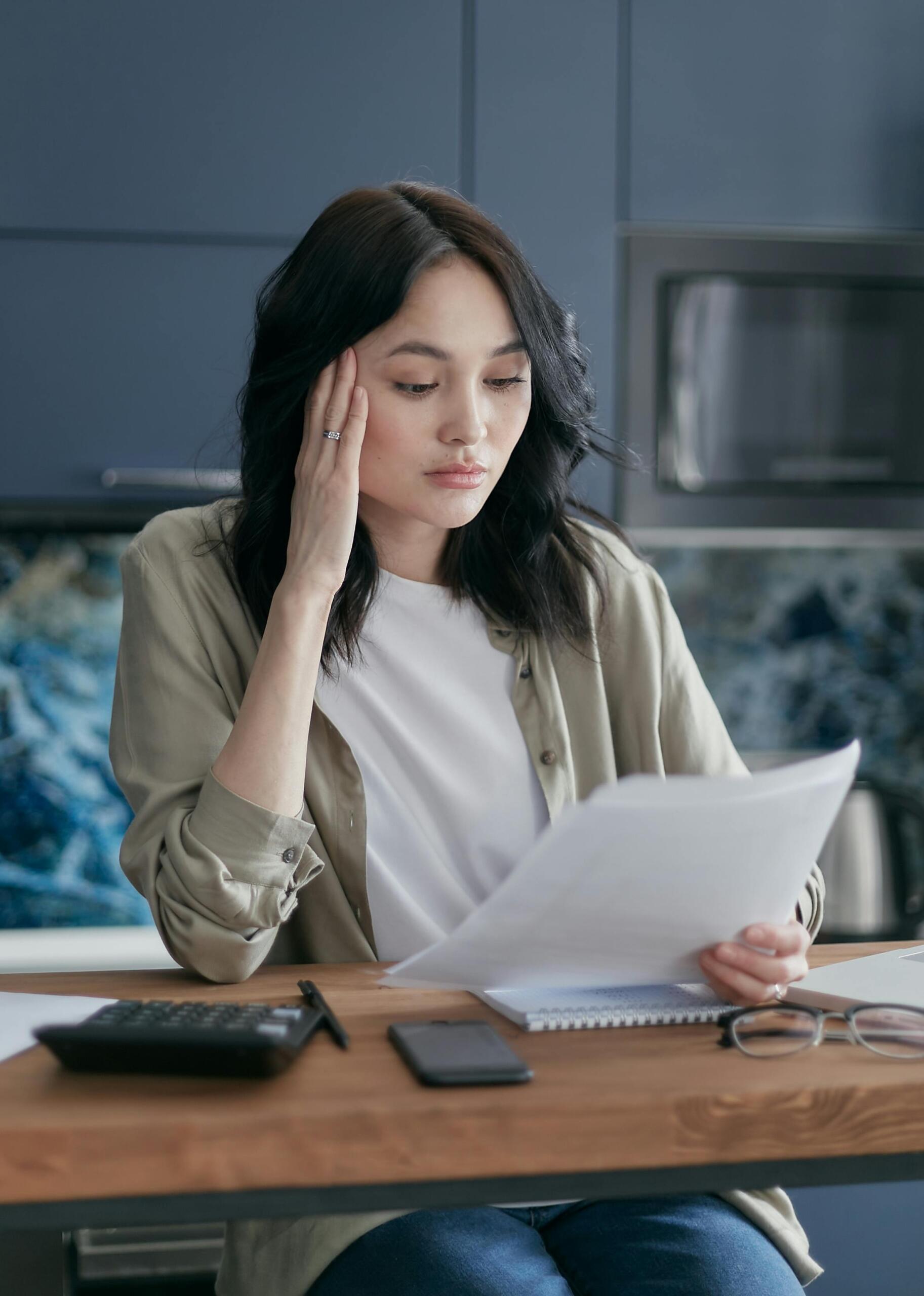 A woman looks thoughtfully at a sheet of paper. Calculators are on her desk. Source: Mikhail Nilov
