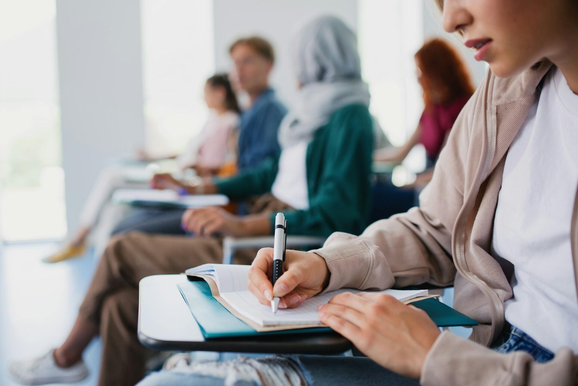 A classroom of college students witting at desks and taking notes with pen and paper