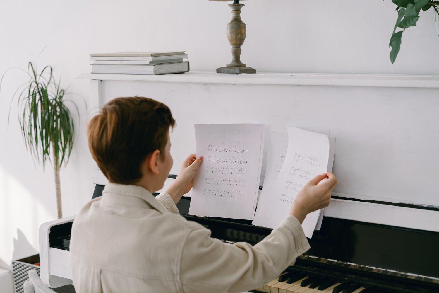 A young person in a light jacket holds sheet music at a piano while a lamp and books sit on a white shelf behind them.