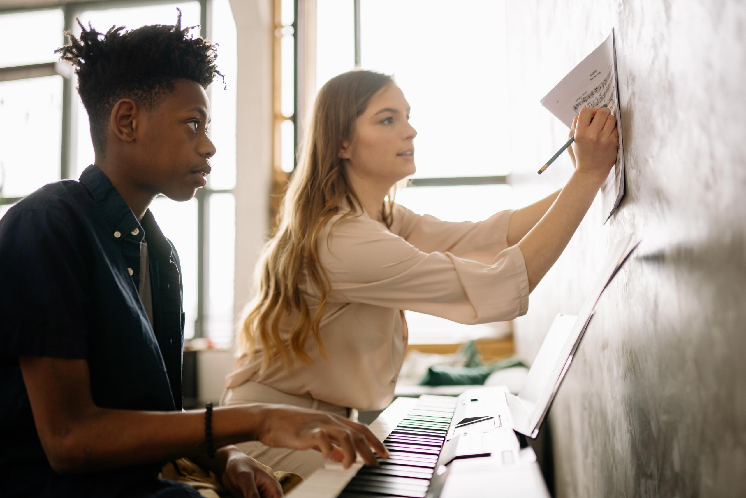 A student plays piano while a teacher writes on a music sheet attached to a wall, with sunlight streaming through a window.