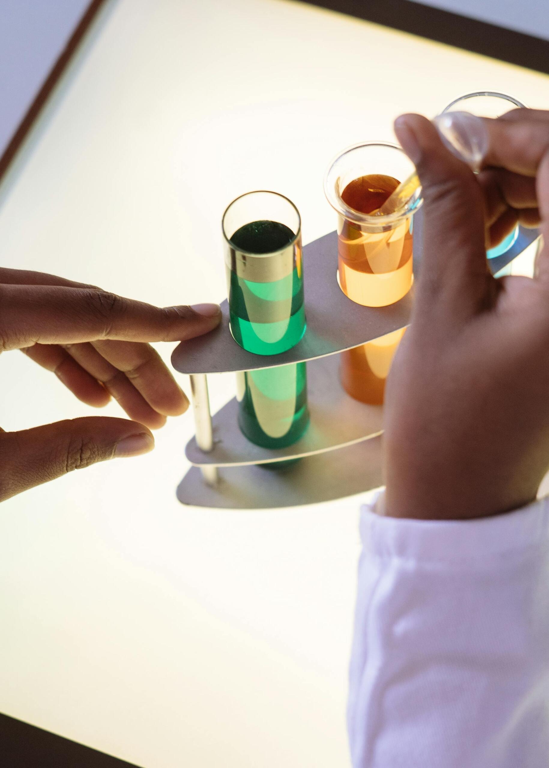 Two hands hold a test tube rack with colorful liquids: green, orange, and blue, under bright light, showcasing a chemistry experiment.