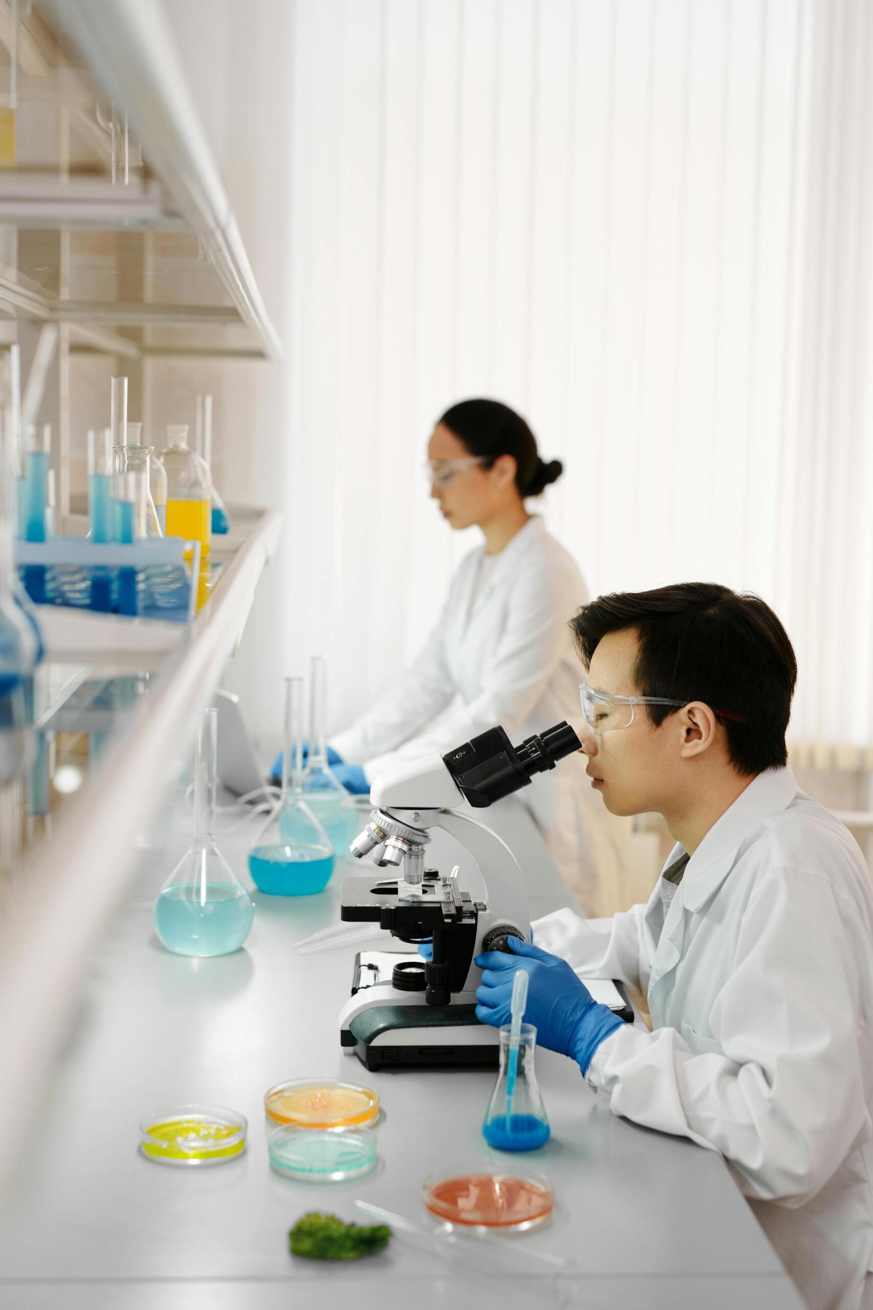 Two scientists in white lab coats work in a bright laboratory, examining colorful liquids through a microscope. Lab equipment is visible.