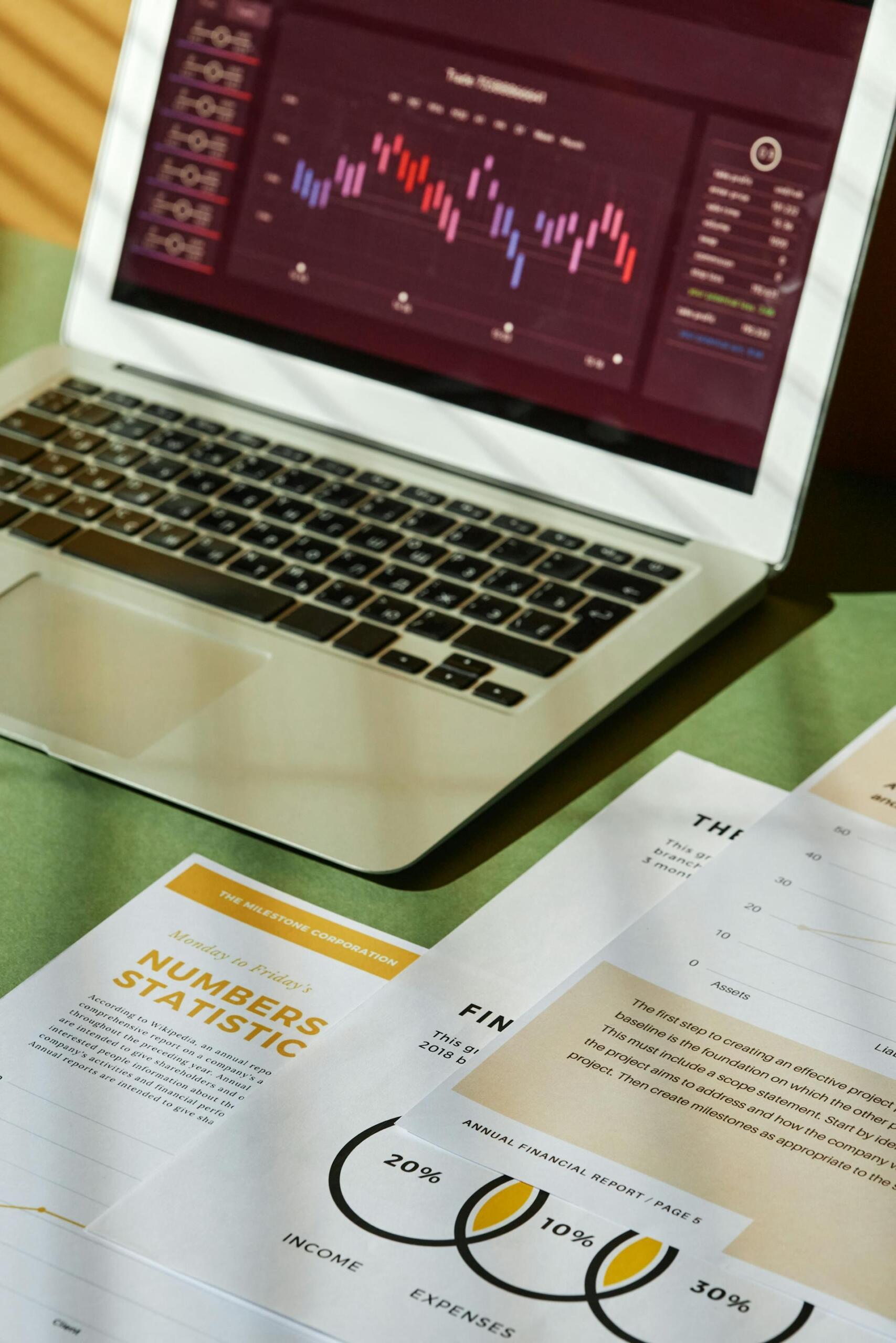 A laptop displays colorful financial graphs, while printed reports on a green table outline a financial analysis project.
