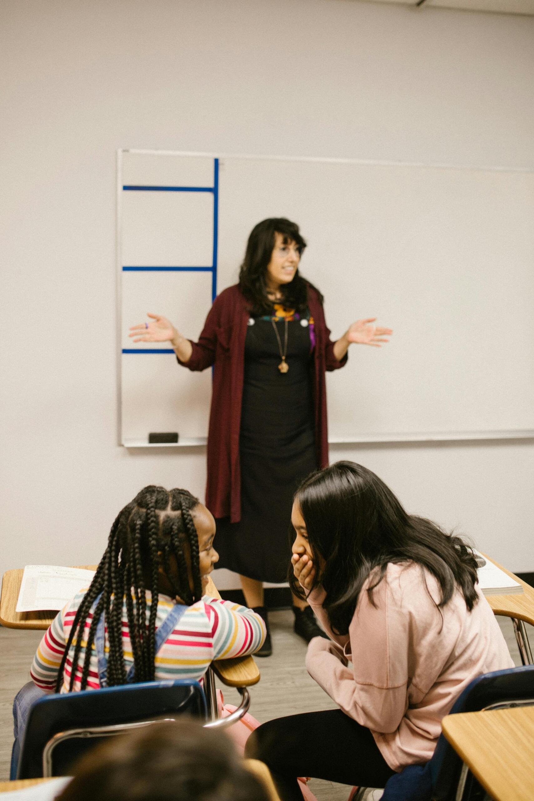 A teacher leads a lesson at the front of the classroom while two students giggle quietly at their desks. Source: RDNE