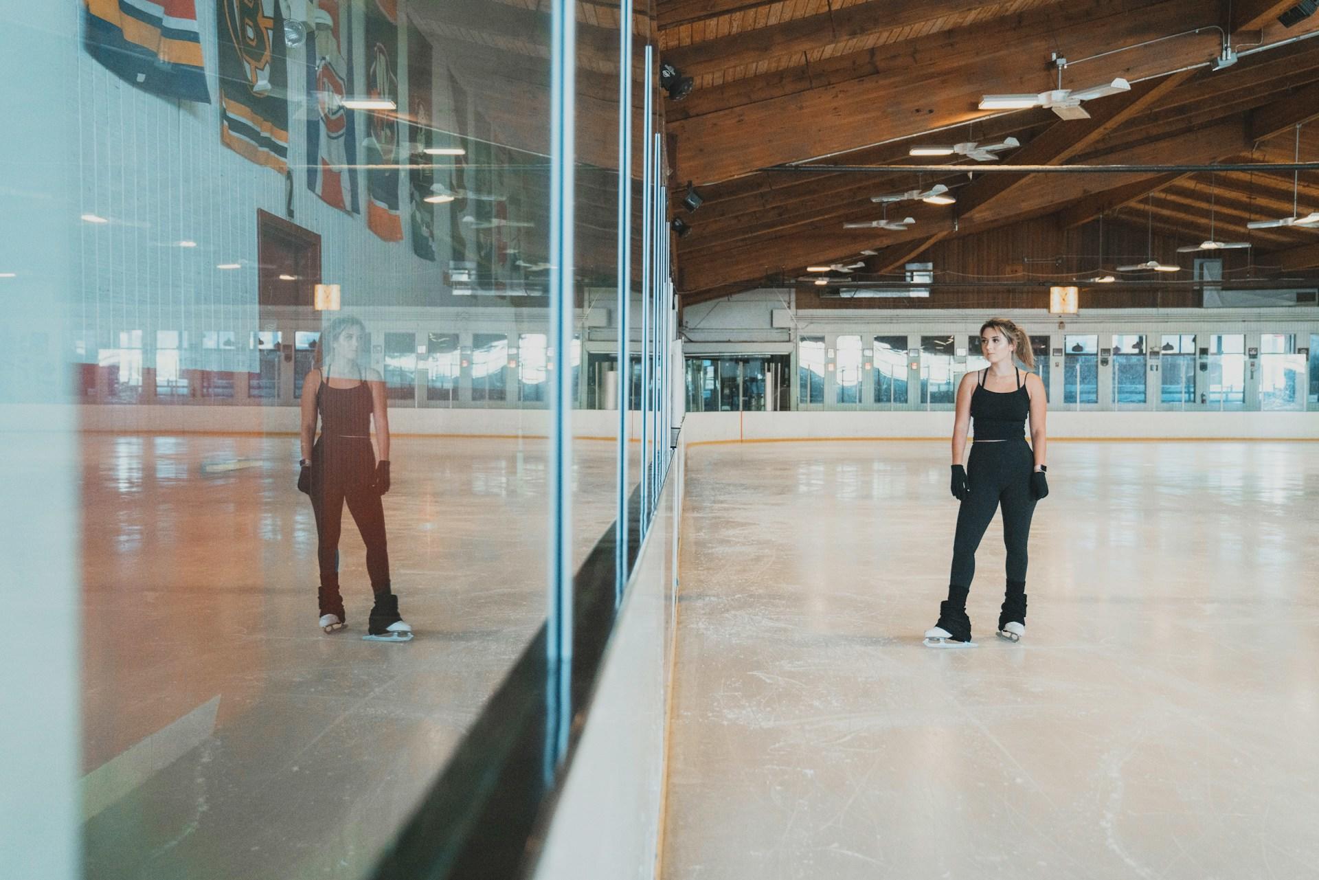 A person in black attire standing on an indoor ice skating rink, reflected in the glass barrier.