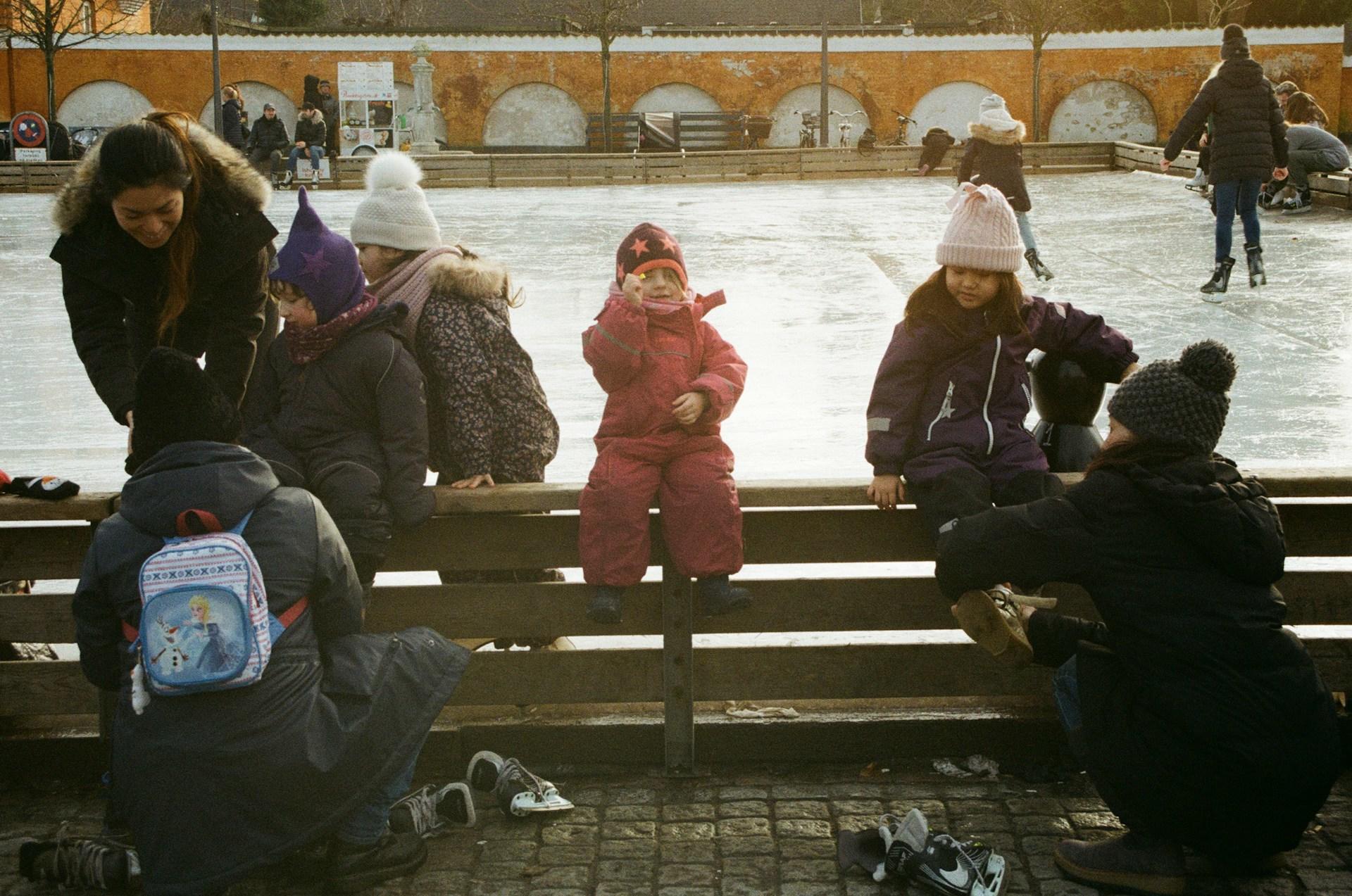 People sitting by an ice rink, with skaters and an old wall in the background.