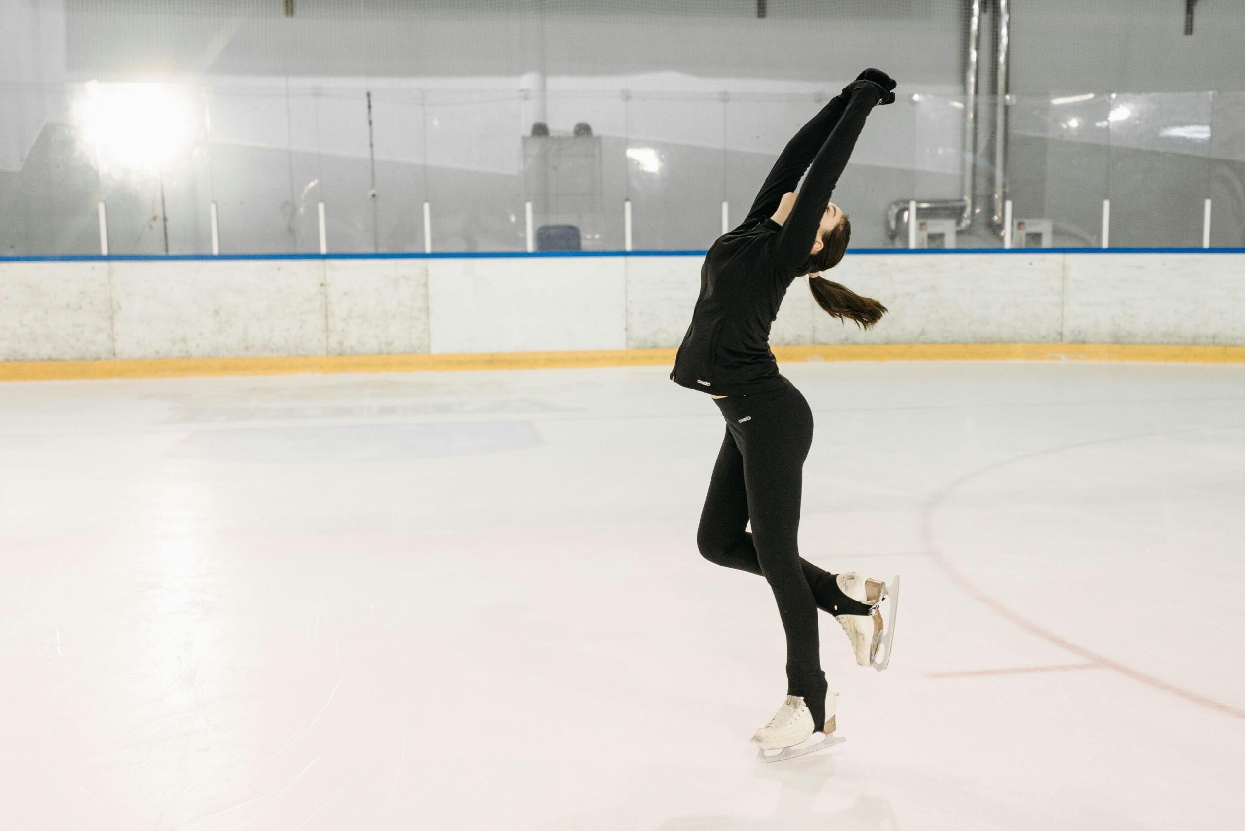 A girl dressed all in black figure skates on an indoor ice rink.