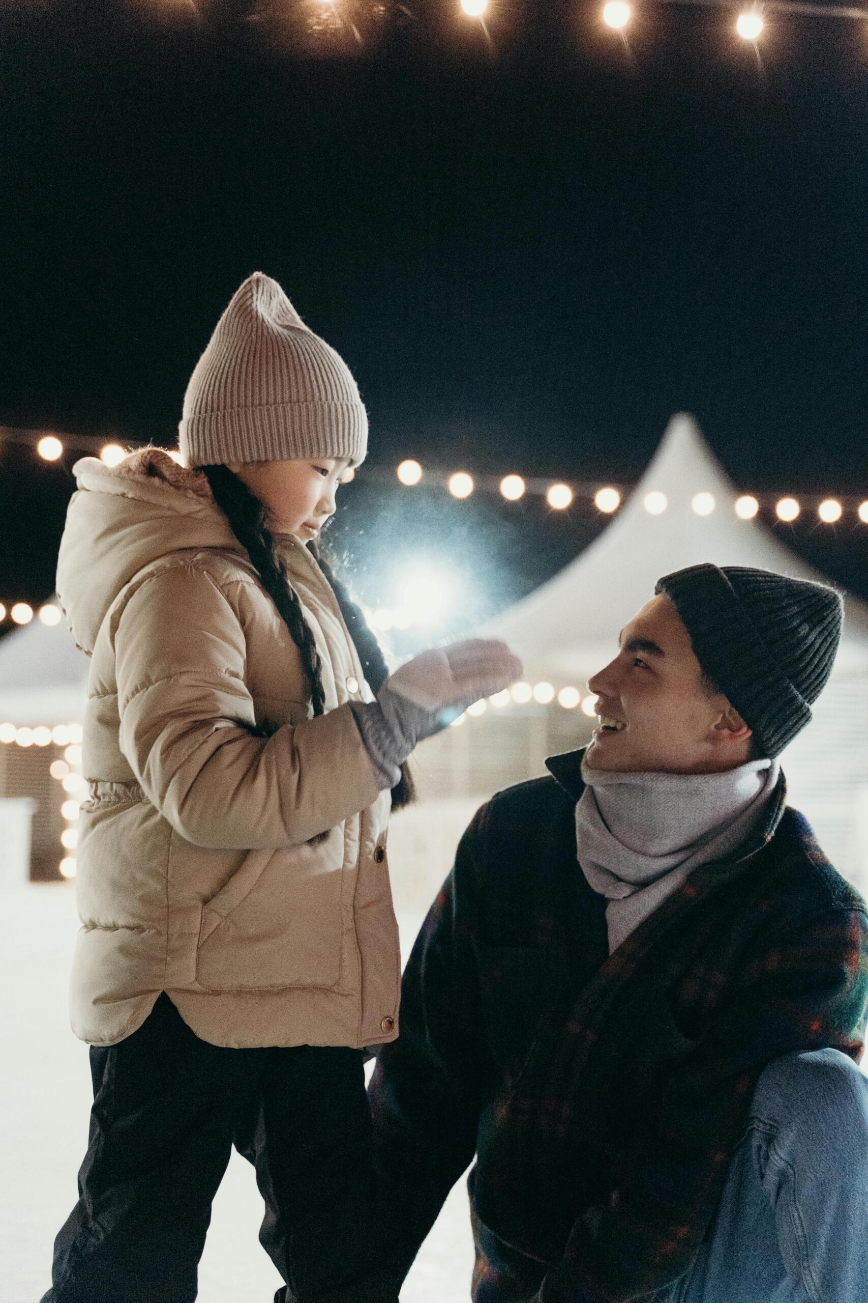 a girl stands on an ice rink with her father. They are dressed in winter attire.
