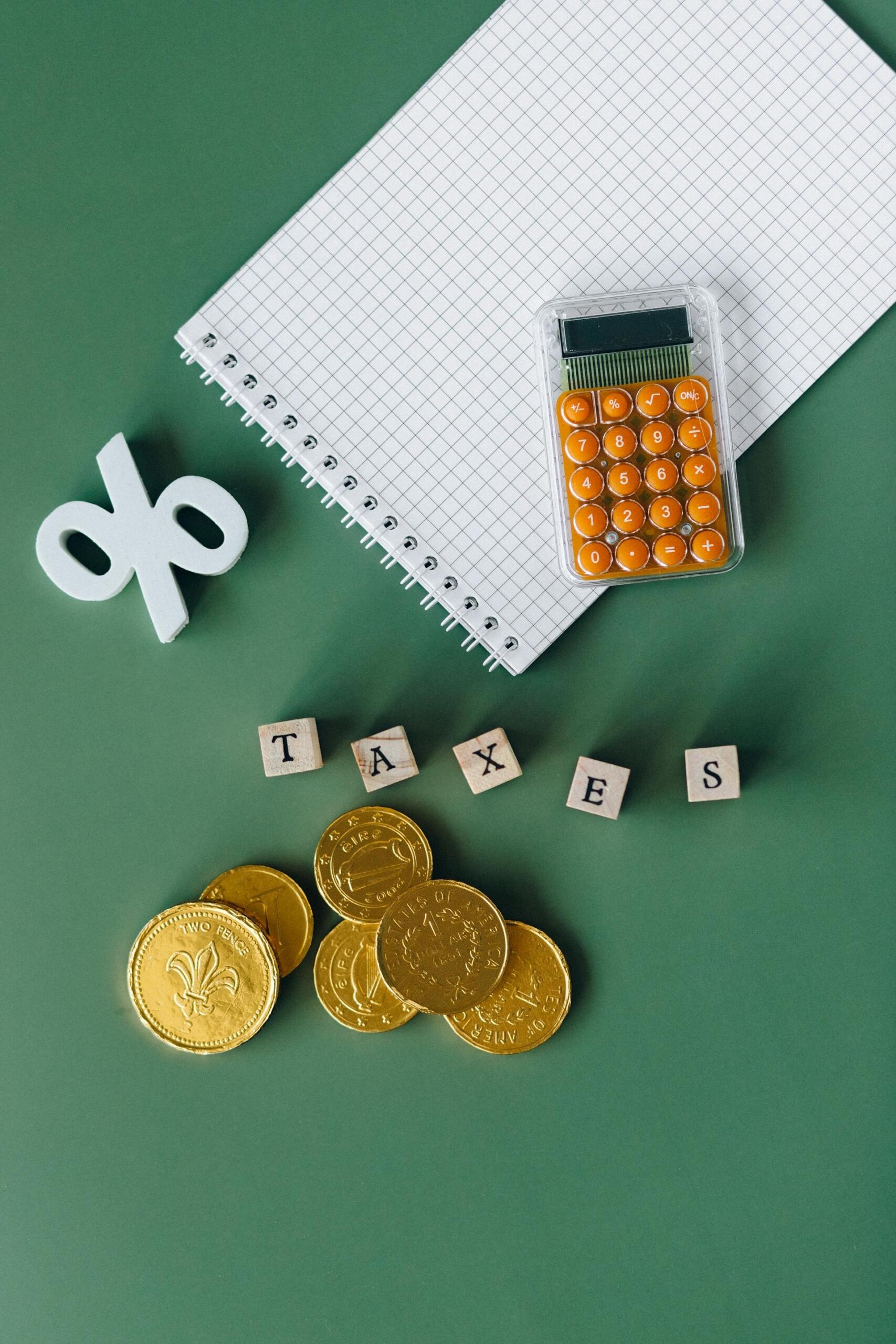 A calculator, notepad, wooden tiles spelling "TAXES," and gold coins on a green background, symbolizing tax preparation.