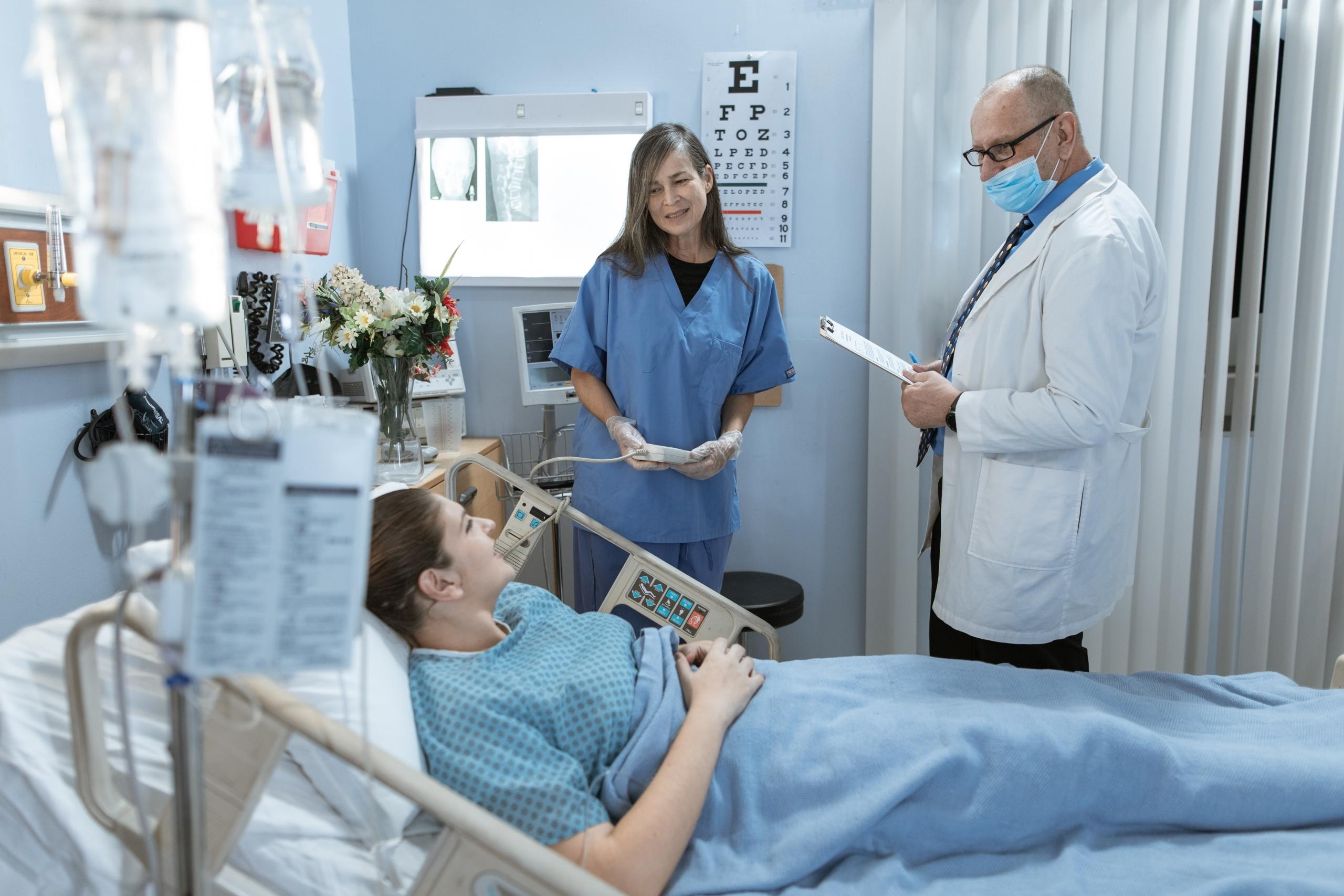Healthcare staff support a patient in a hospital bed, there are some eye charts and x-rays in the background.