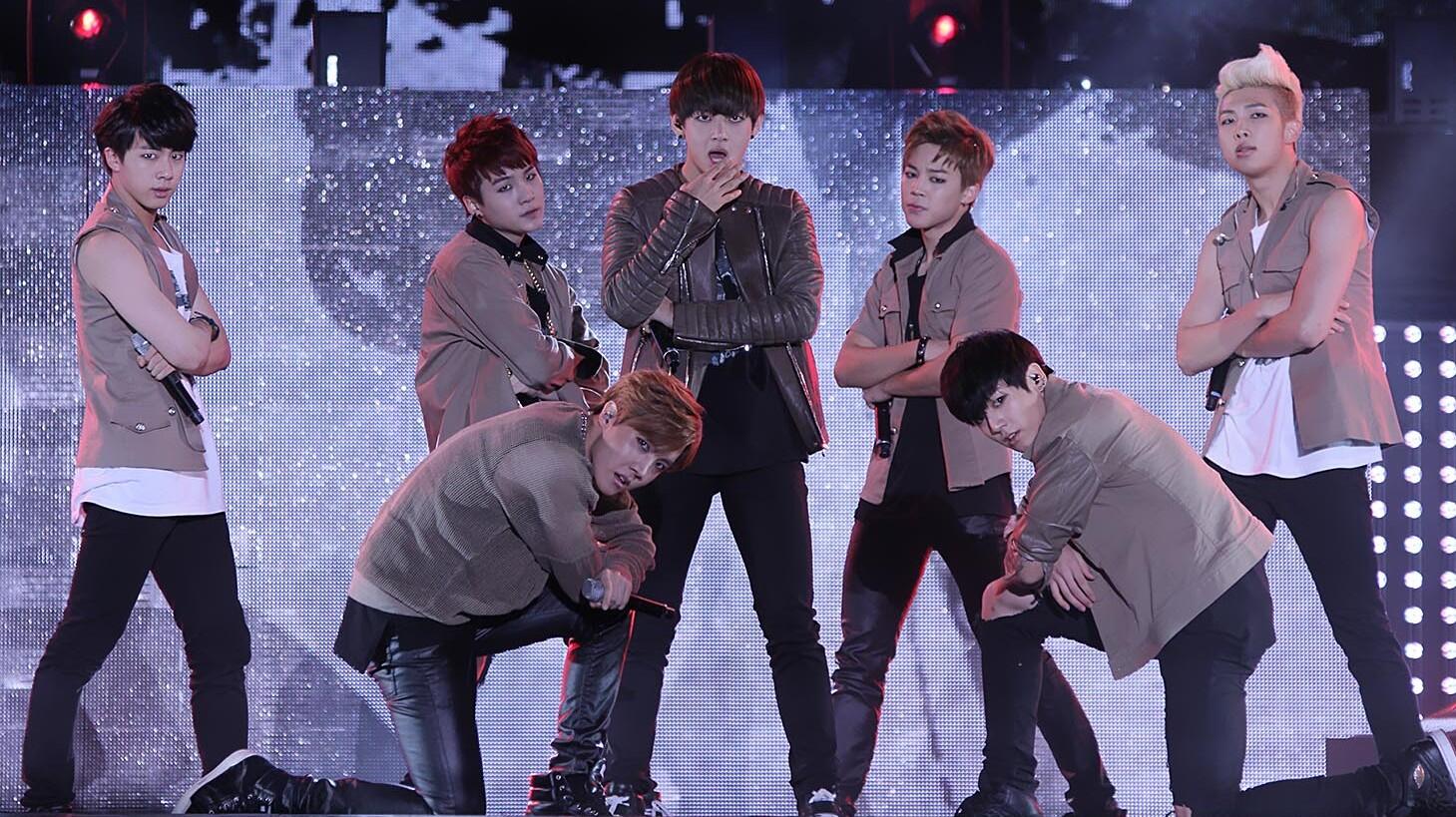 A group of seven male performers dressed in stylish stage outfits singing and posing under bright lights during a live concert, with a vibrant blue-lit background behind them.