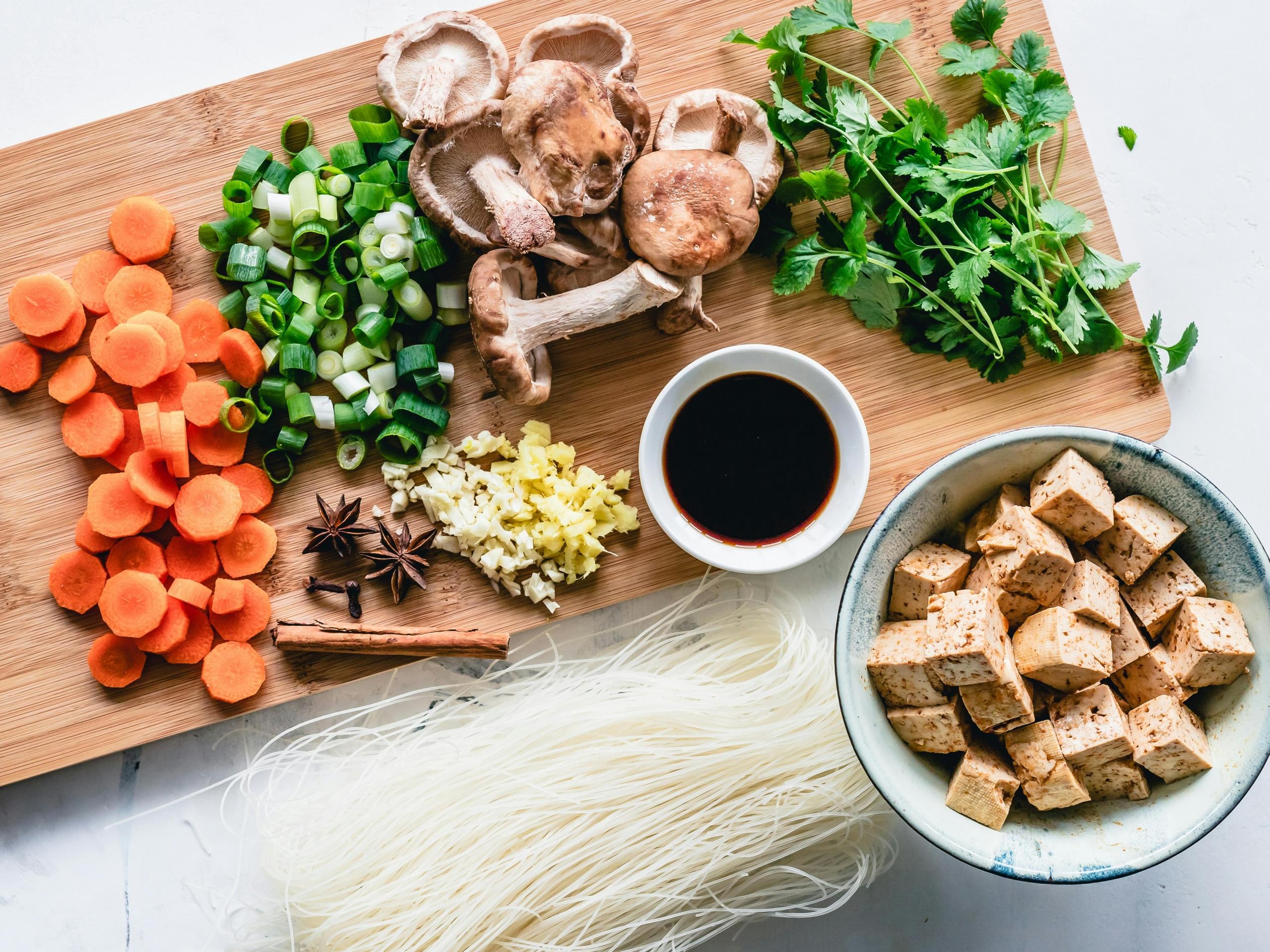 A cutting board that has carrots, green onion, tofu, garlic and mushrooms all cut up on top.