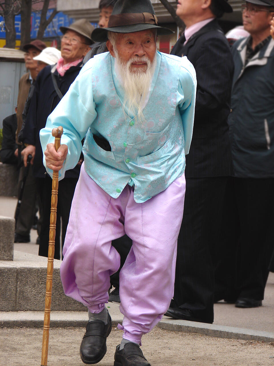 An elderly Korean man stands gracefully in the street, wearing a traditional hanbok that blends soft pastel tones.