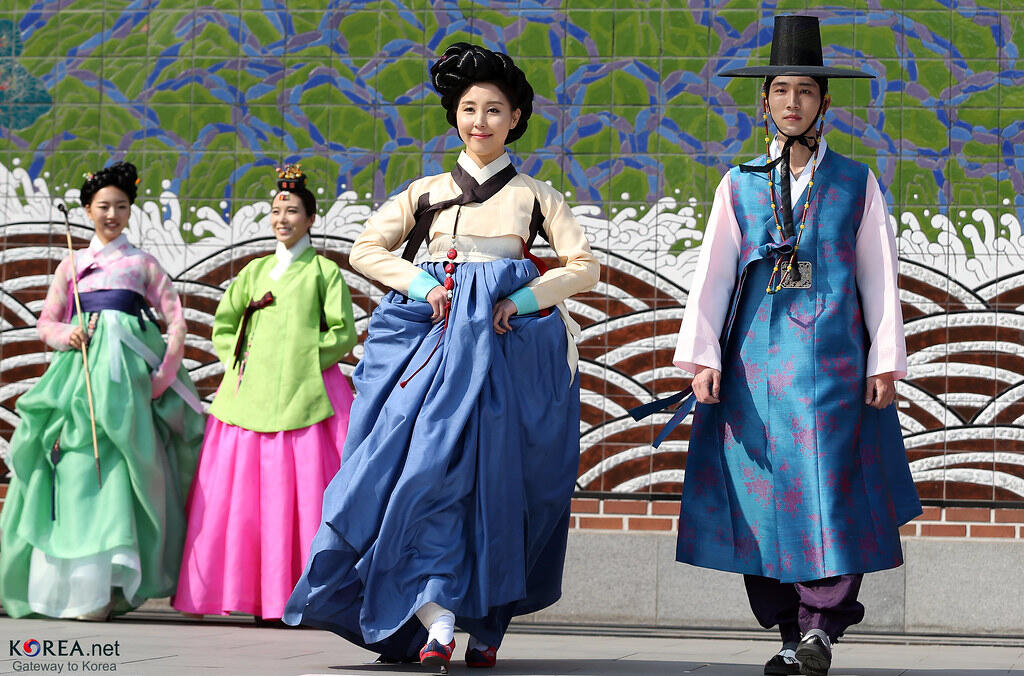 Four individuals in traditional Korean hanbok attire walk in front of a colorful mural depicting waves and mountains.