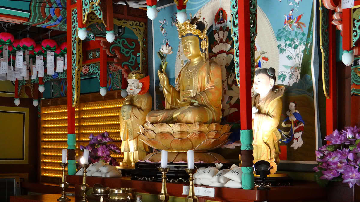 Inside a serene Korean Buddhist temple, a golden statue of Buddha sits gracefully upon a lotus pedestal, radiating calm and enlightenment. The Buddha holds a white lotus flower, symbolizing purity and spiritual awakening.
