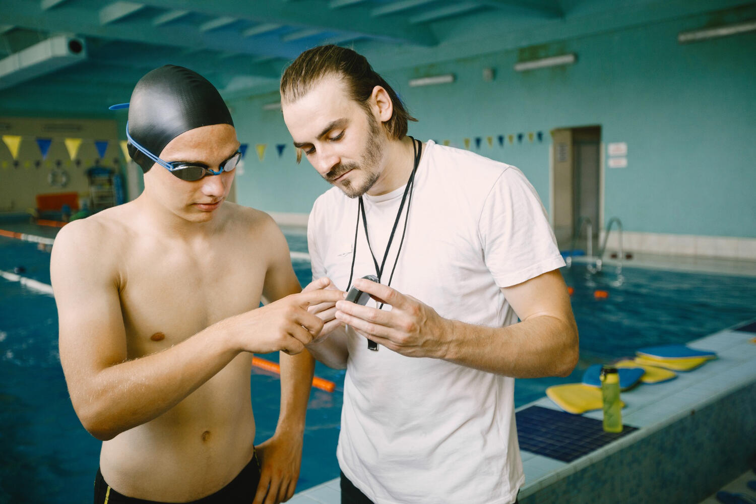 A swimming coach and a student discuss training techniques at a pool, wearing swim gear and focused on a small device.