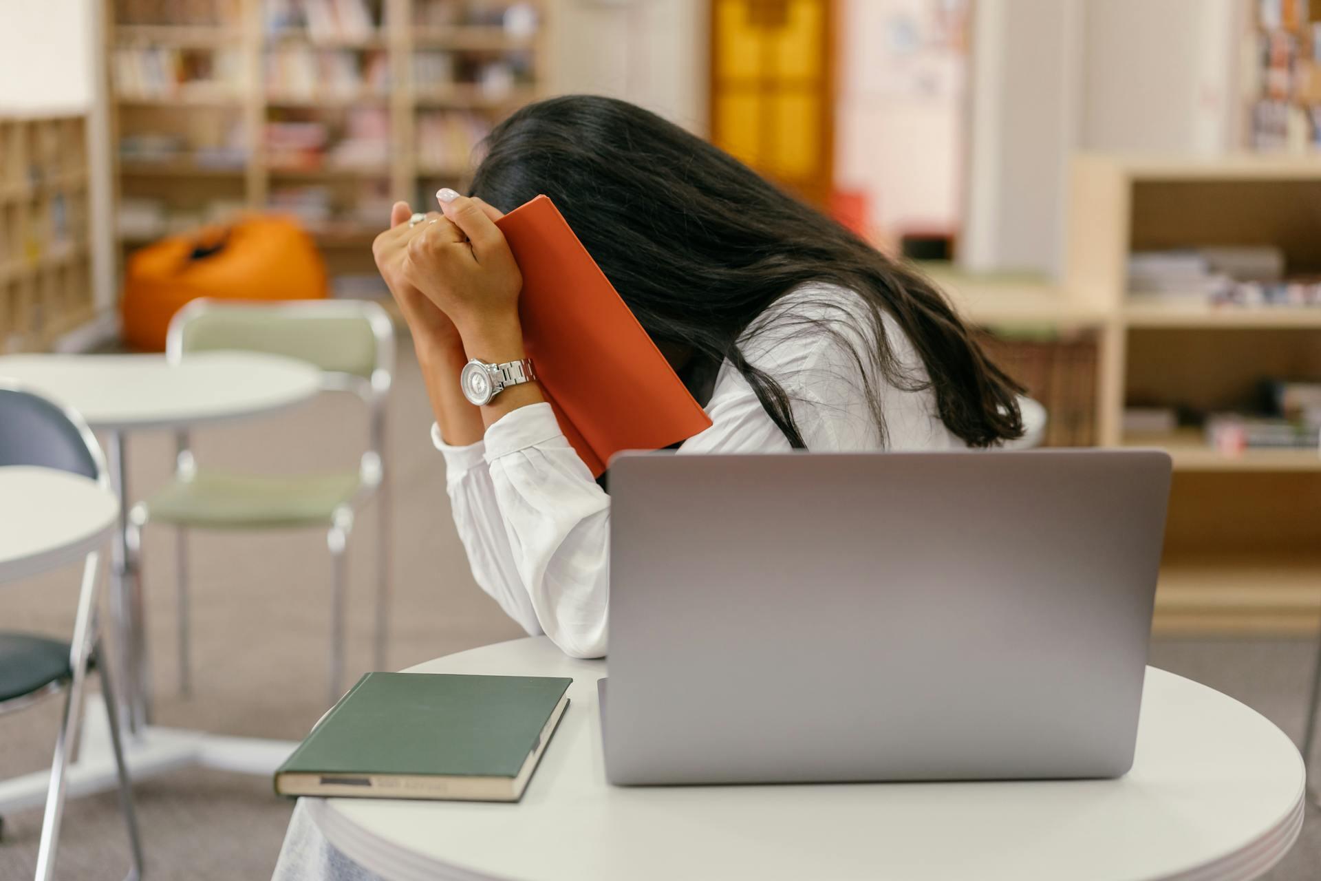 A student sits at a table in a library, covering their face with a book, visibly stressed, with a laptop and another book nearby.
