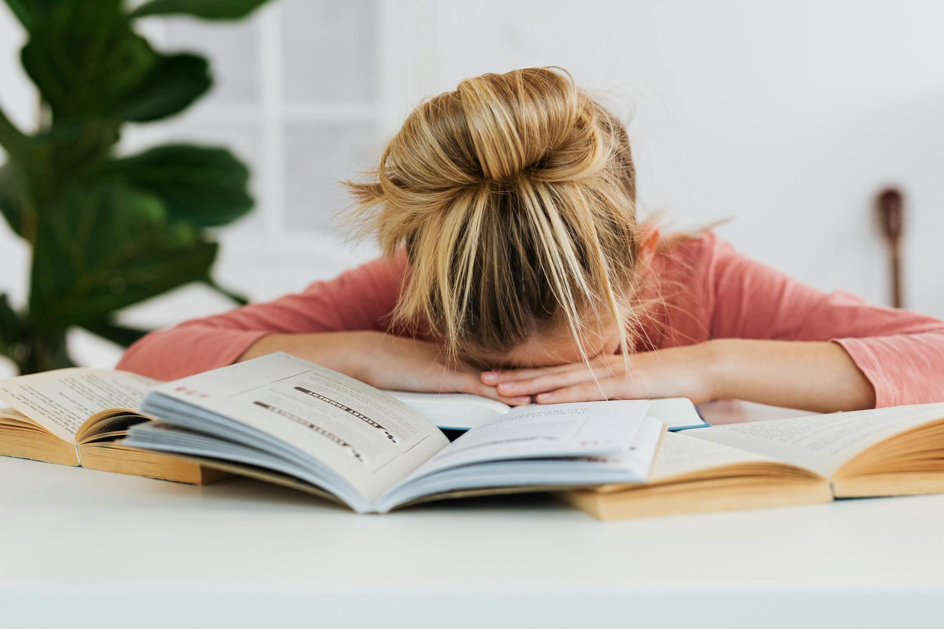 A tired student rests her head on an open book, surrounded by several other books, in a brightly lit study space.